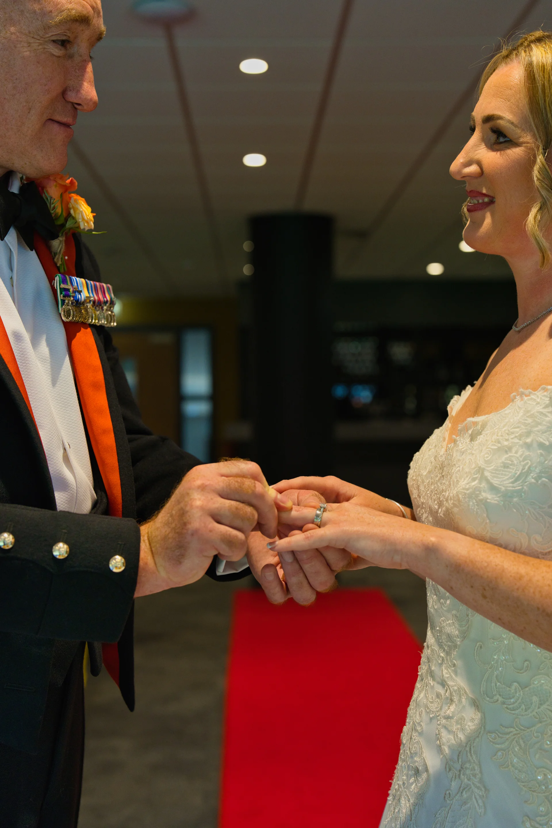 A groom in a military uniform with medals exchanges wedding rings with a smiling bride wearing a lace gown.