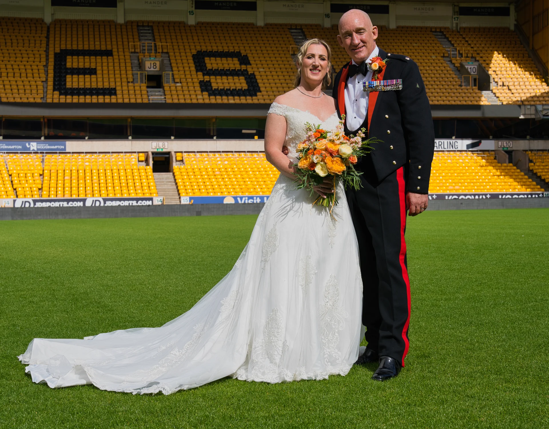 A newlywed couple poses on the pitch of a vibrant yellow sports stadium, with the bride in an off-the-shoulder lace gown featuring a long train and holding an orange bouquet, and the groom in a ceremonial military uniform.
