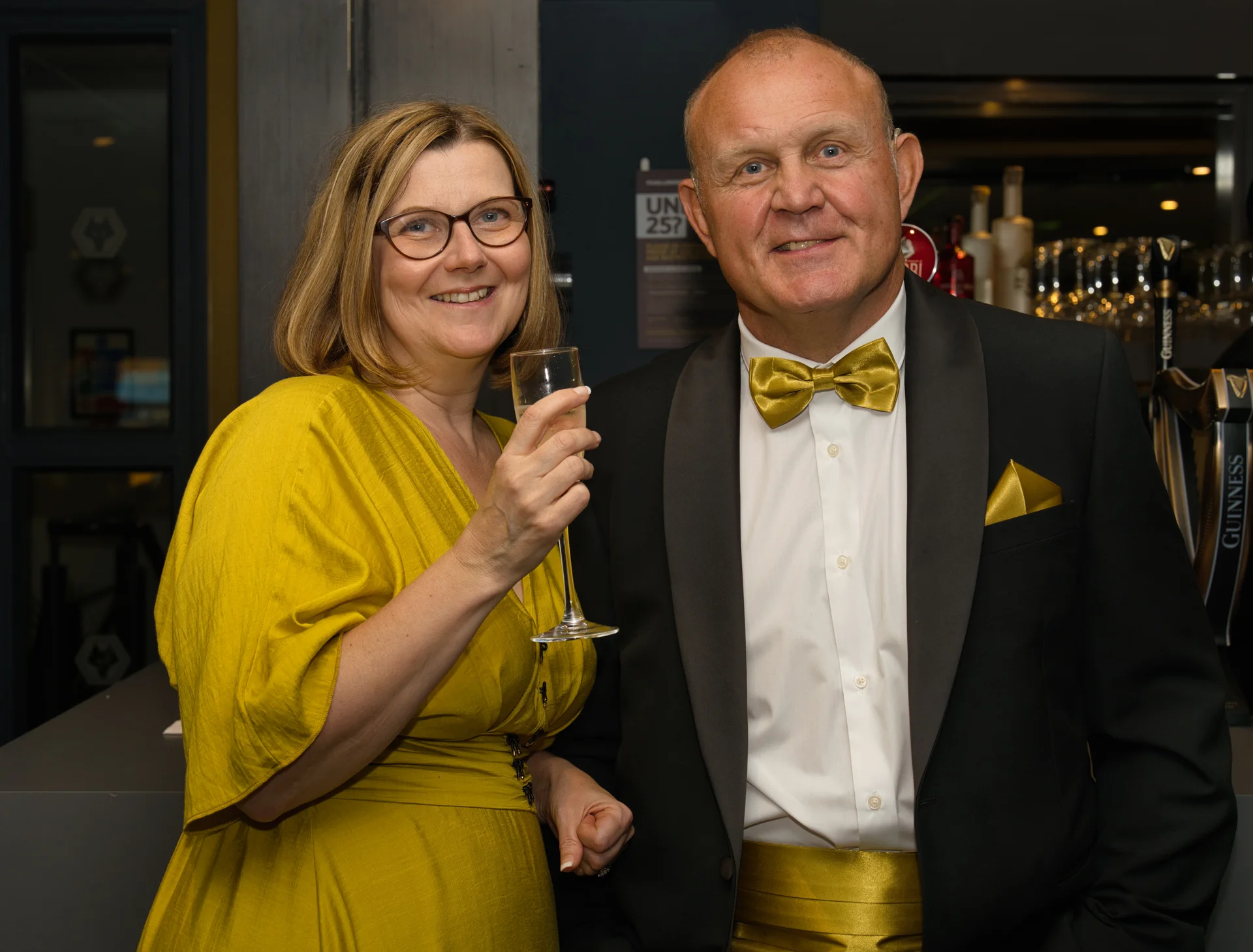 An older couple smiling together at a wedding evening reception, with the guest holding a champagne glass and wearing a black tuxedo with gold accessories.