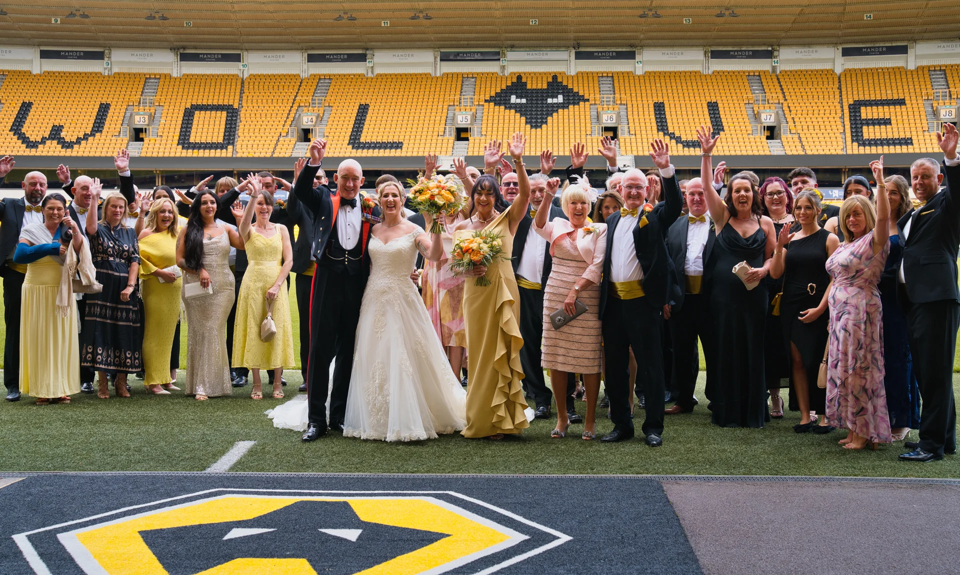 A newlywed couple and their large group of wedding guests cheer with raised hands on the pitch of a yellow and black stadium.