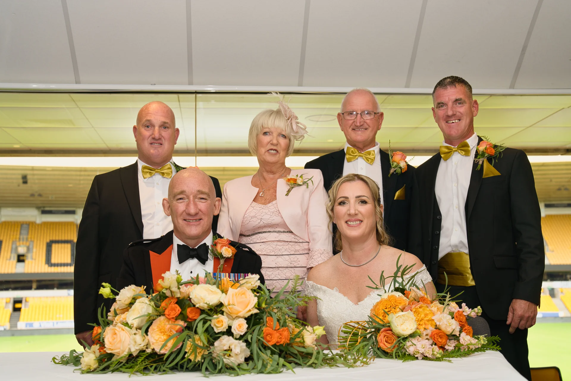 A smiling wedding party including the bride and groom, mother of the bride, and groomsmen posed together at the top table decorated with vibrant orange floral arrangements.