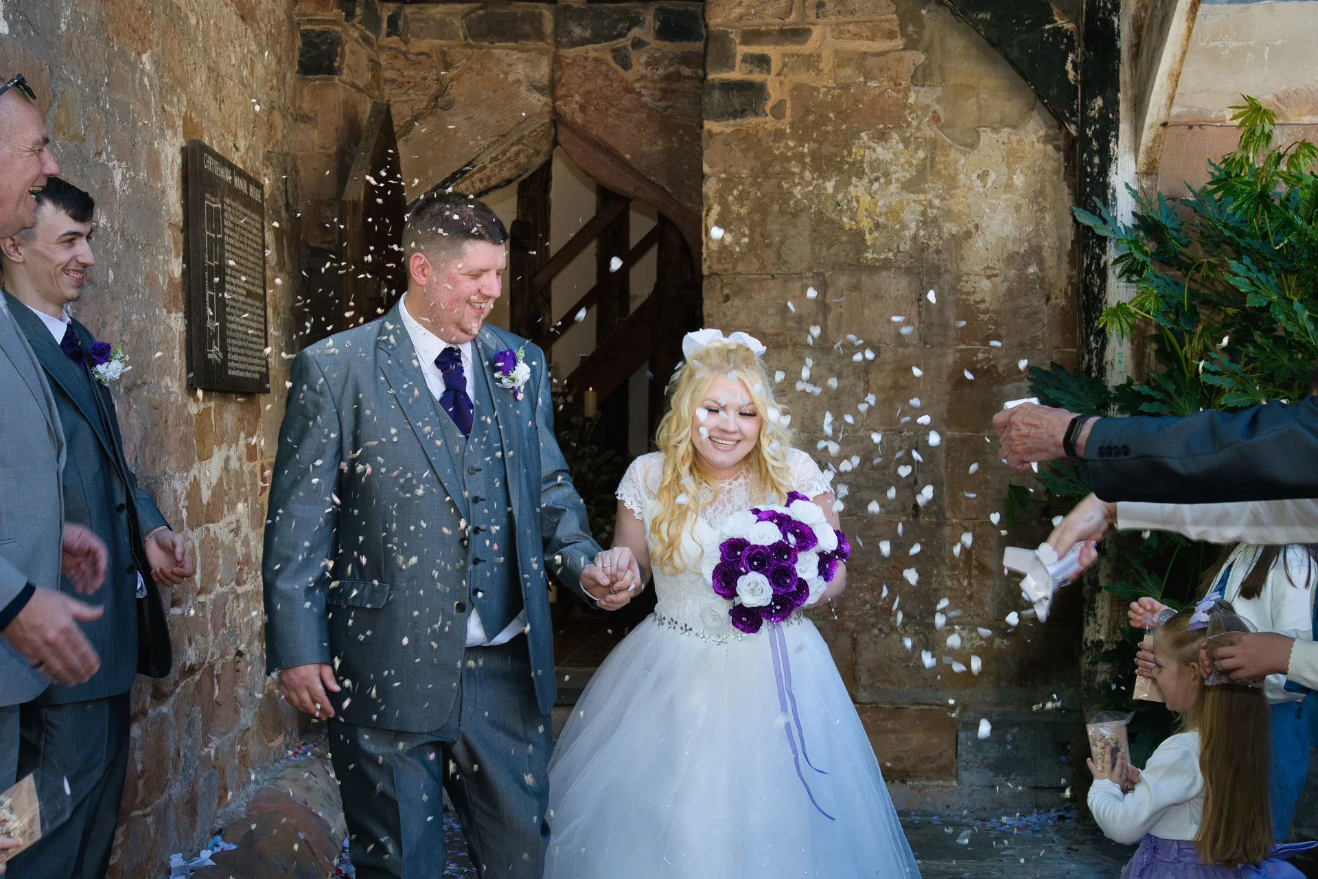 A newlywed couple walk hand in hand through a shower of confetti outside a historic stone doorway while guests throw petals.