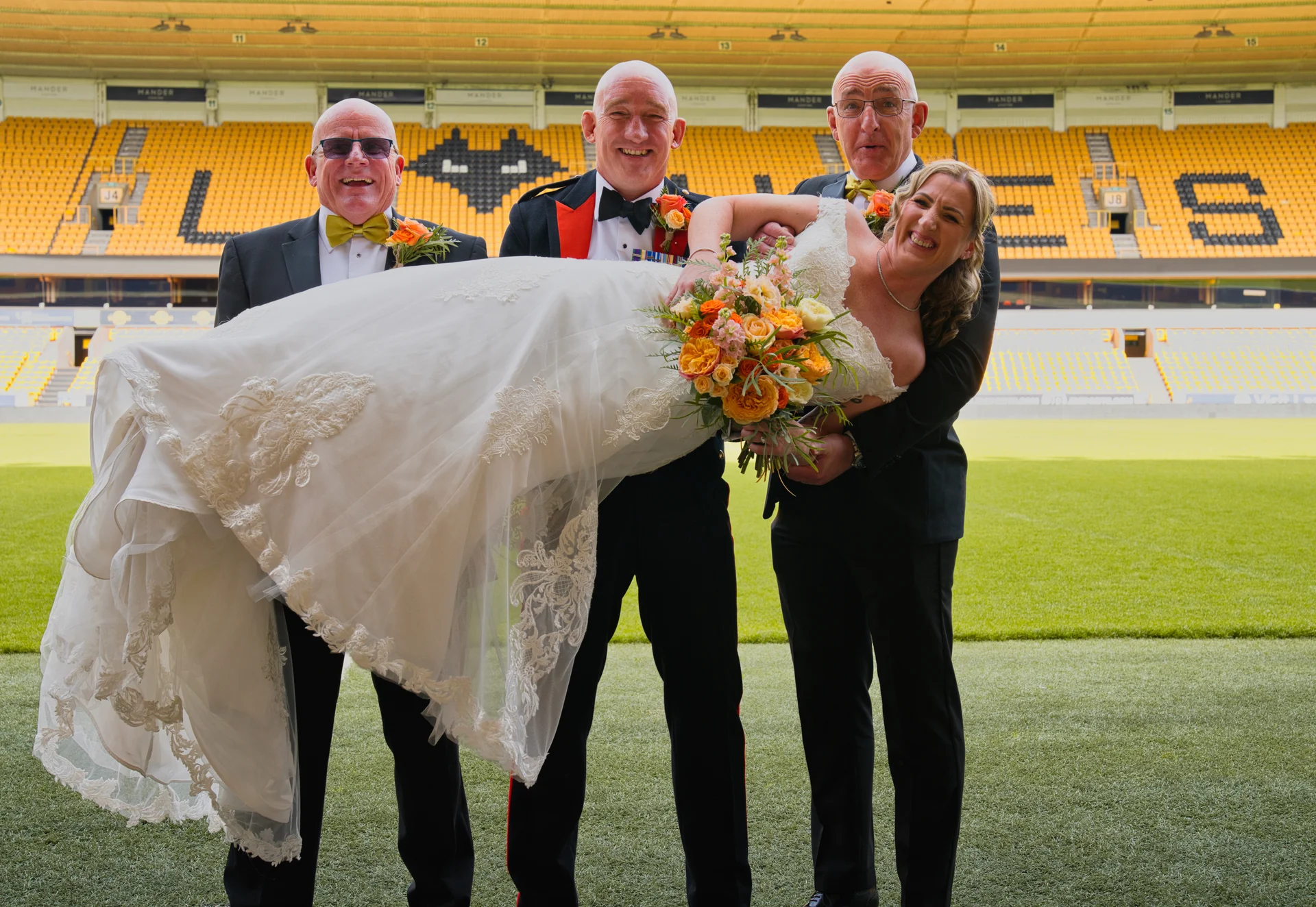 Three smiling men playfully carry a laughing bride horizontally across the pitch of a large football stadium.