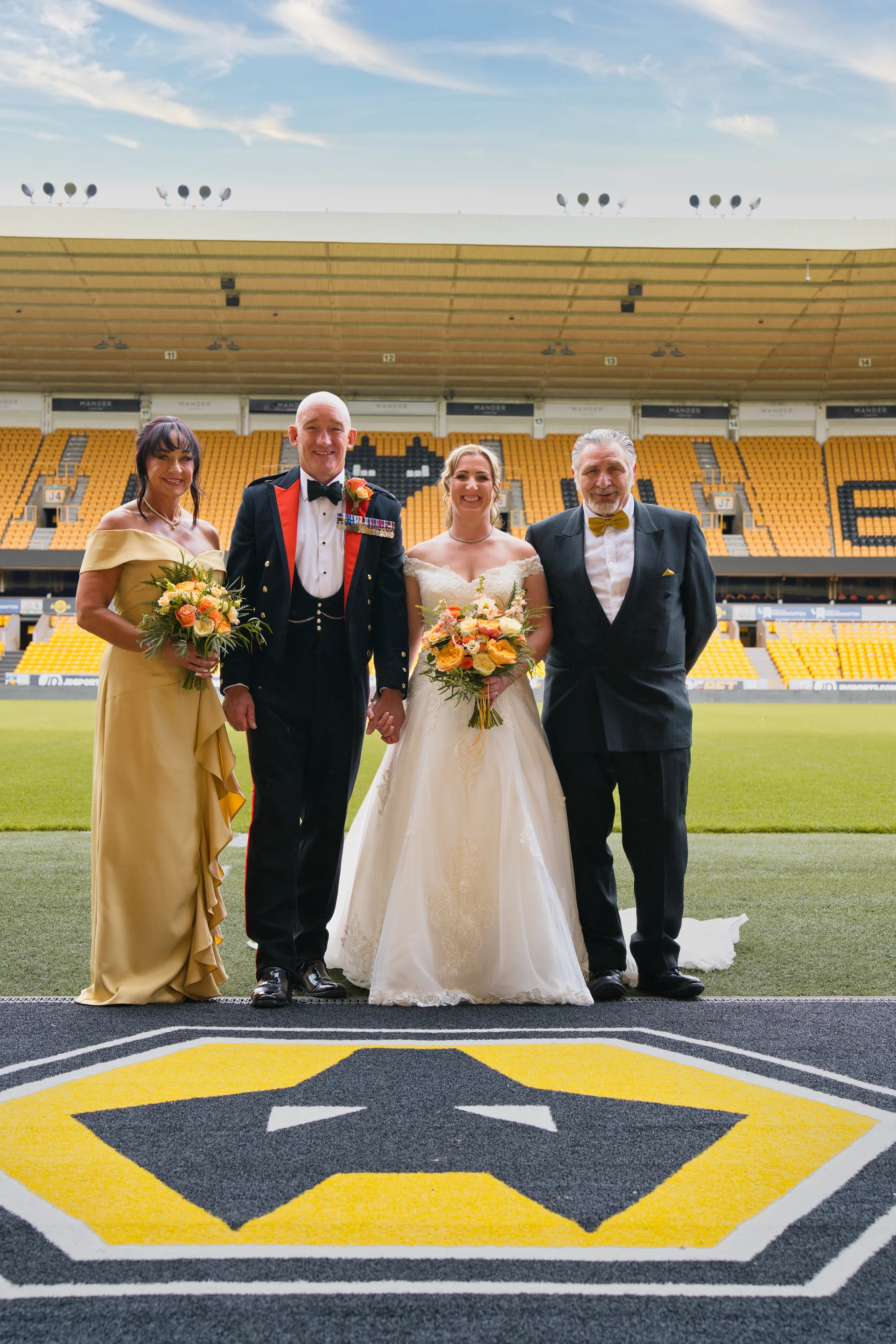 A newlywed couple and two family members pose for a formal group photograph on the pitch at a sports stadium.