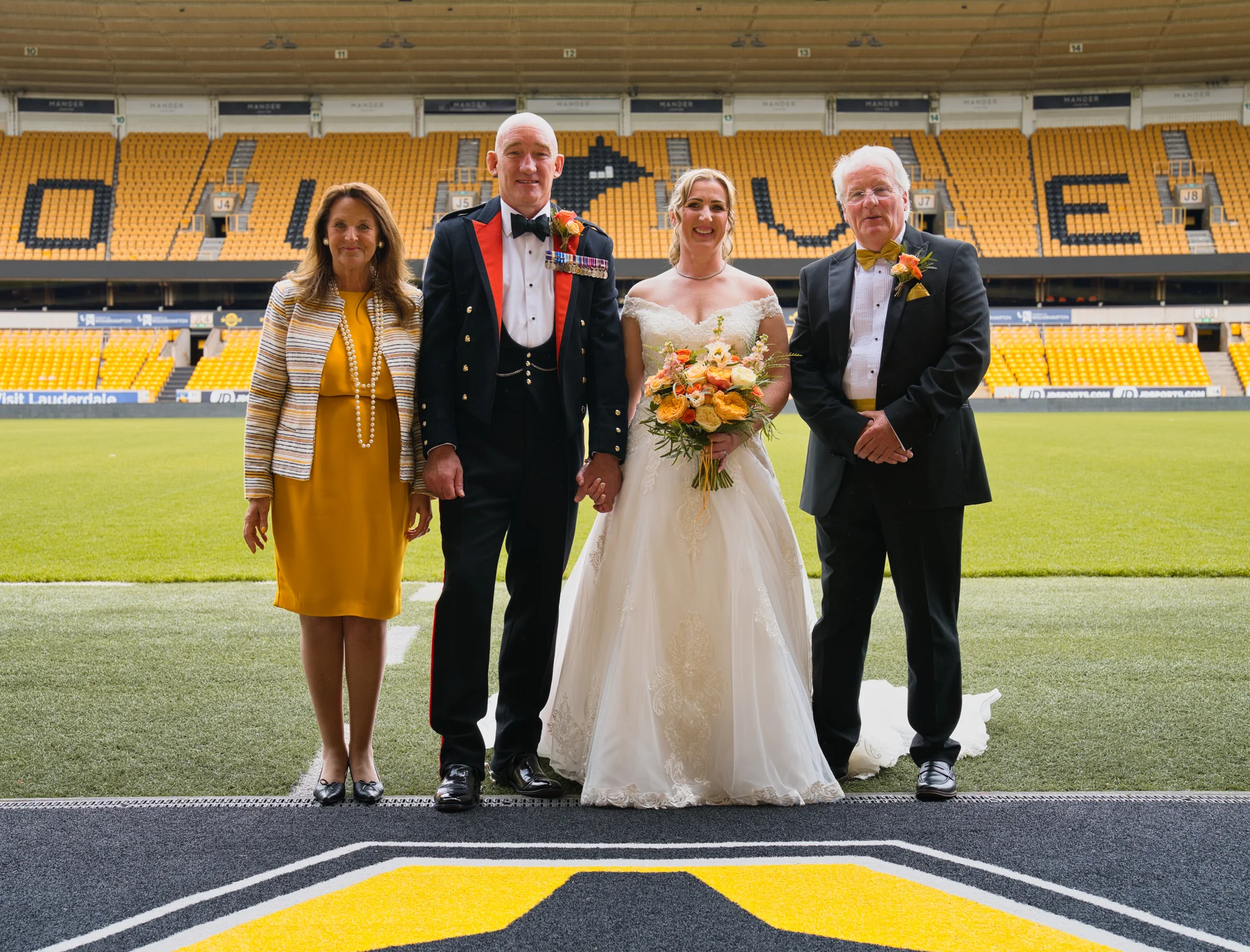 A newly married couple in full wedding attire pose with their parents on the pitch of a large football stadium with yellow seating.
