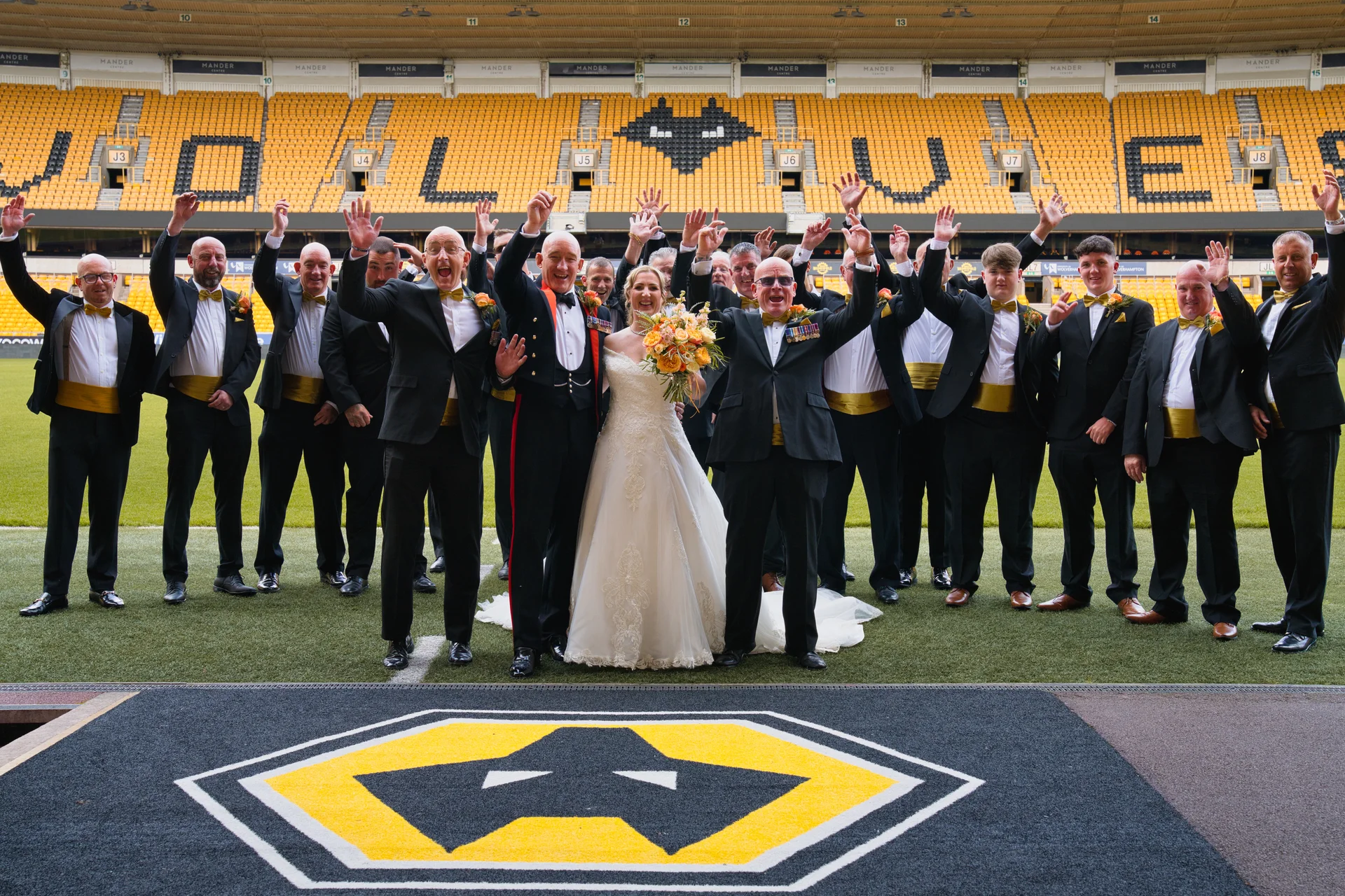A newlywed couple and a large group of groomsmen in black tie attire cheer with their hands raised on the pitch of a football stadium.