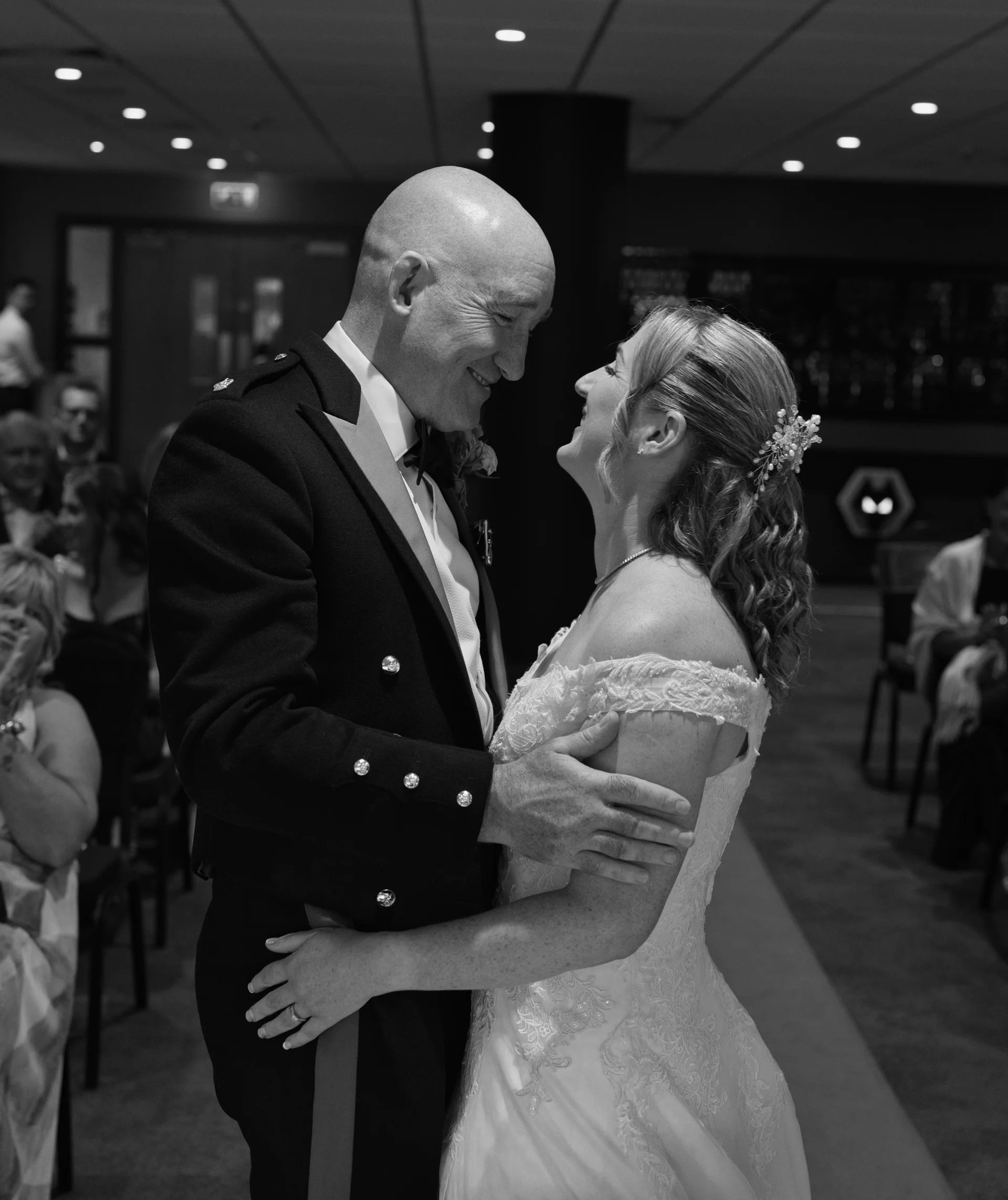 An older bride and groom share a tender, smiling embrace during their indoor wedding celebration captured in a classic black and white style.