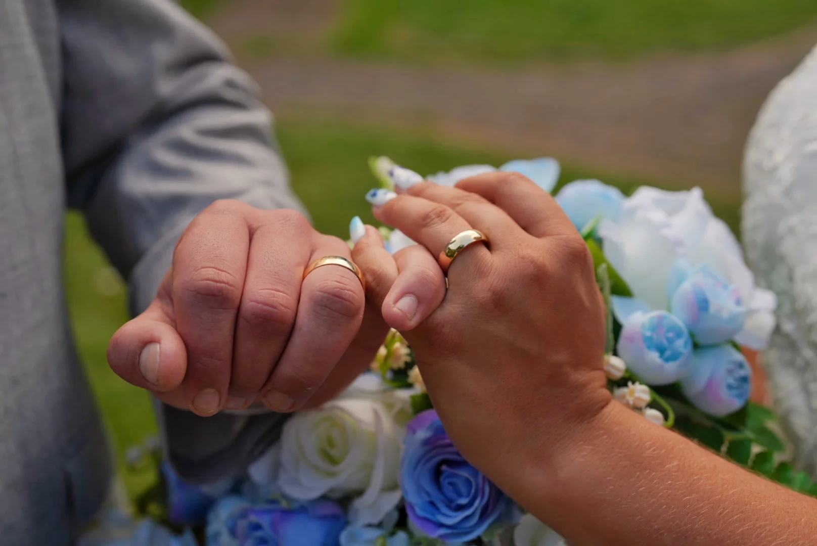 Close-up of a newly married couple holding hands to proudly display their shiny gold wedding bands in front of a blue and white floral bouquet.