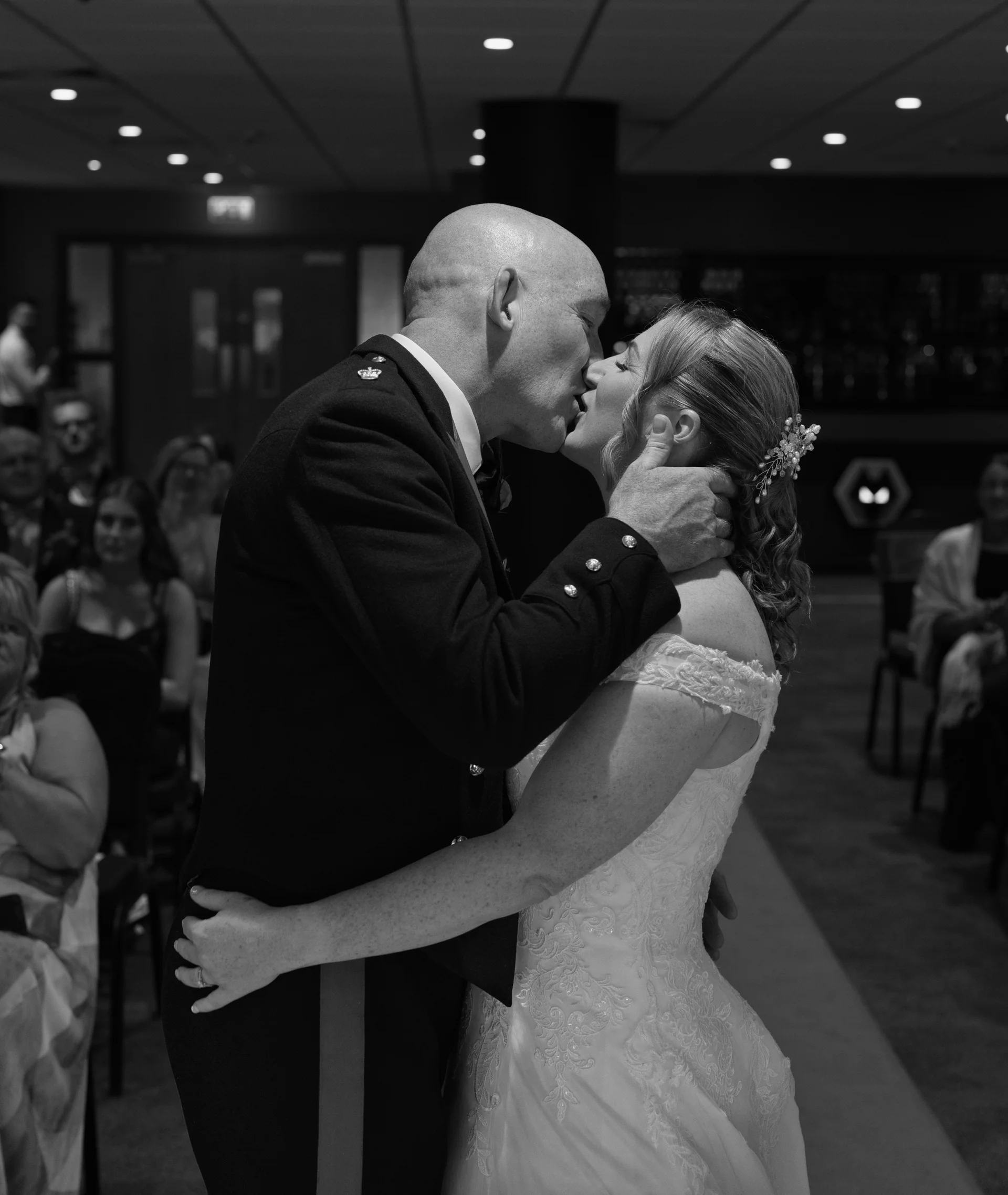 A newlywed couple shares their first kiss during an indoor civil ceremony, with the groom in a military uniform and the bride in an off-the-shoulder lace dress.