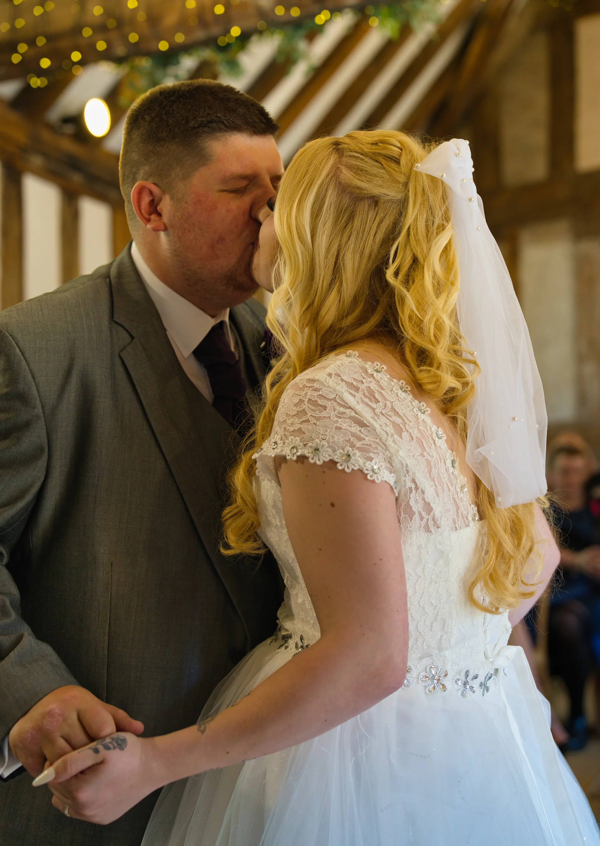 A newly married couple share their first kiss inside a rustic oak-framed barn with exposed rafters and warm lighting.