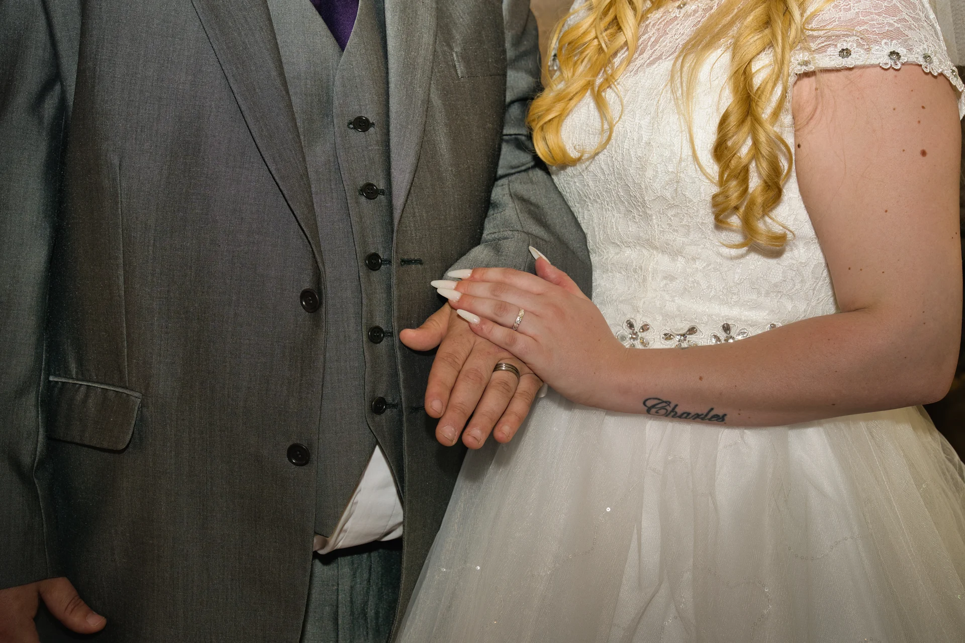 A close-up shot showing a newly married couple holding hands, highlighting their wedding rings and a wrist tattoo.