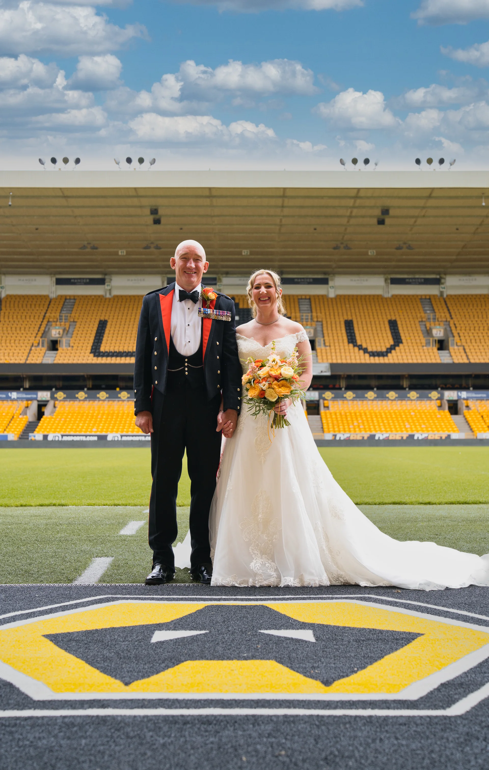 A smiling bride and groom standing pitchside inside a large yellow and black football stadium, with the bride holding a colourful hand-tied floral bouquet and wearing an off-the-shoulder gown while the groom wears a military mess dress uniform.