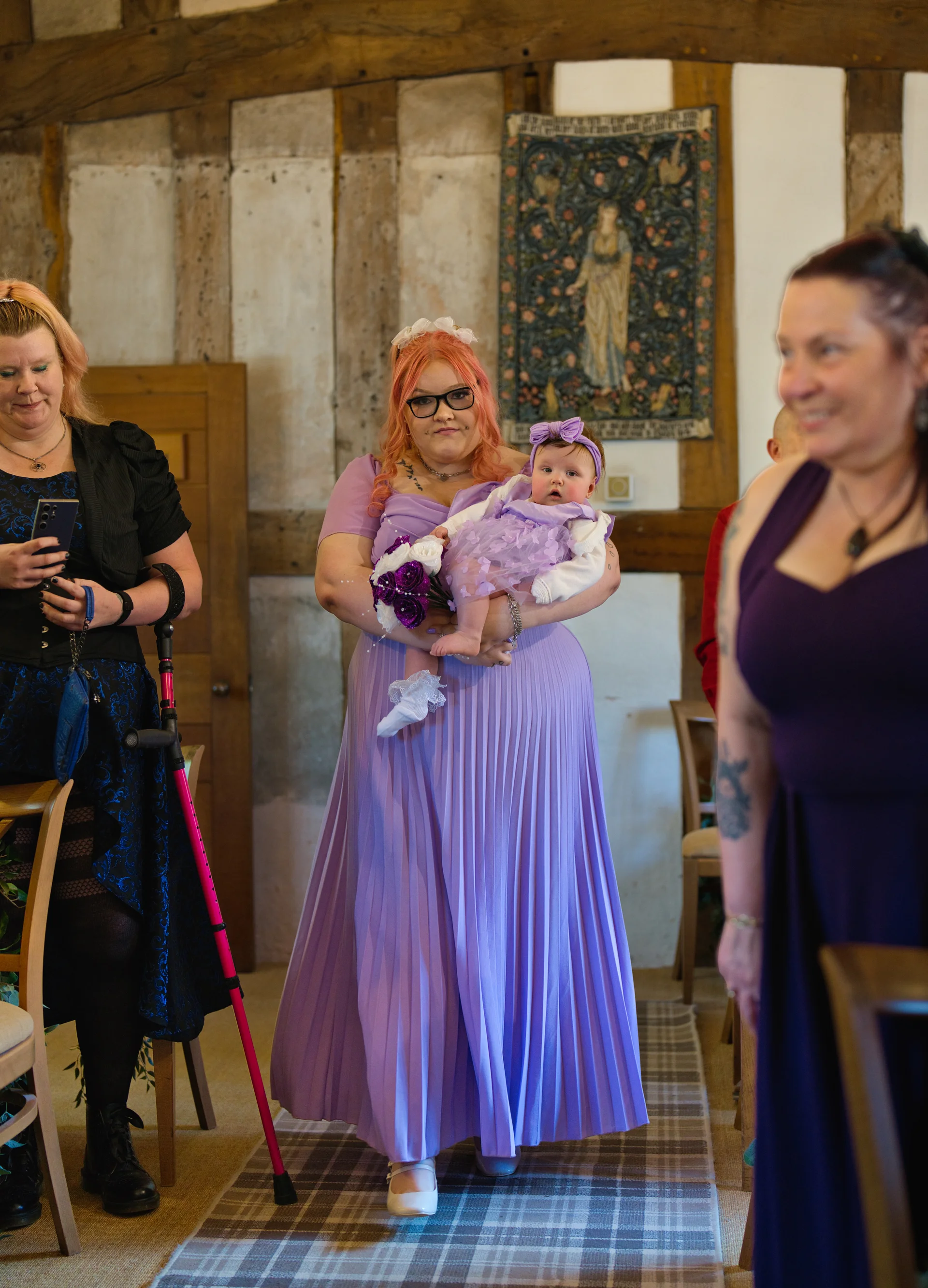 A guest holding a baby and a bouquet walks down the aisle inside a rustic barn with exposed wooden beams, surrounded by fellow guests.