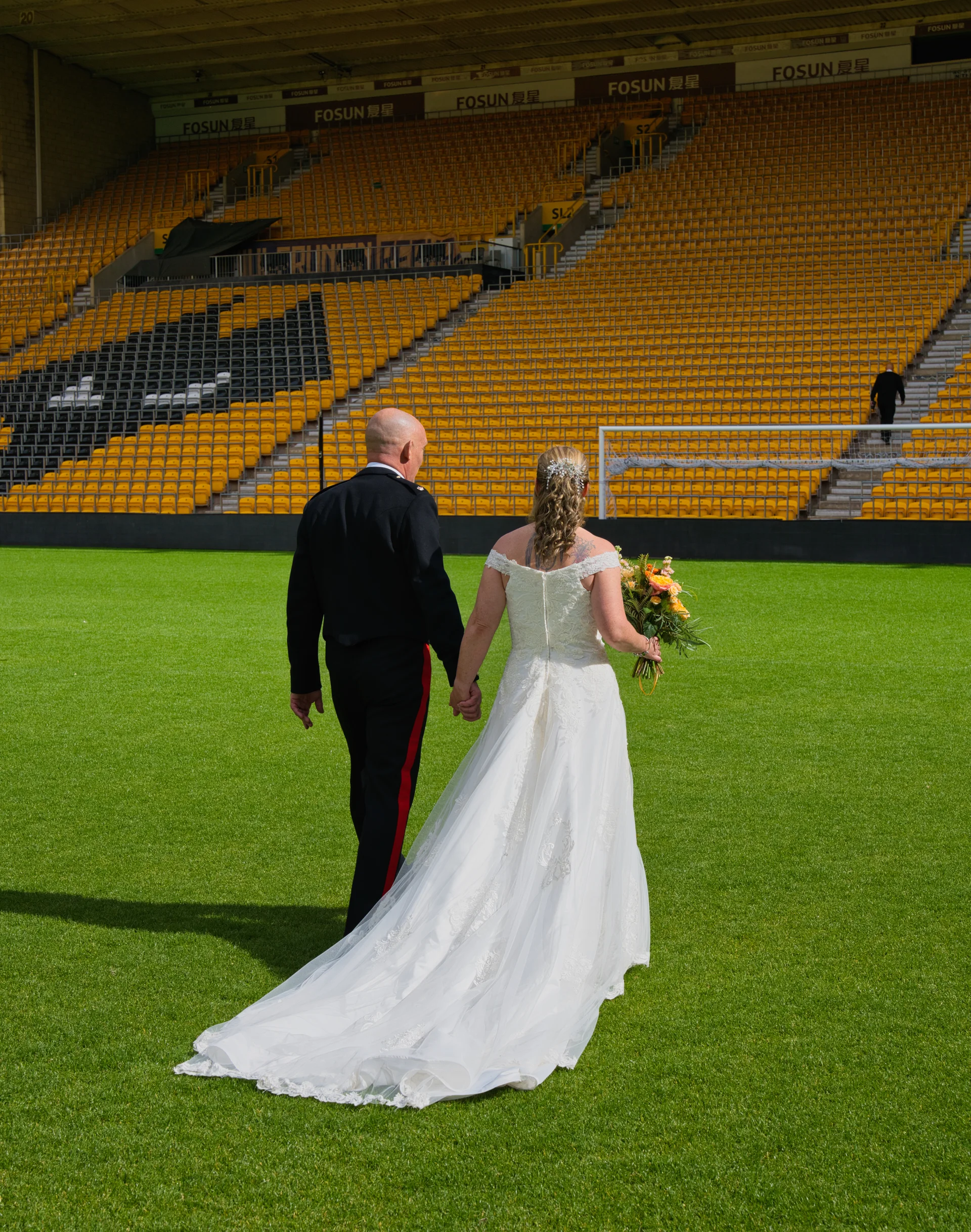 A newlywed couple walks hand in hand across the vibrant green pitch of a large yellow-seated sports stadium, with the bride wearing an off-the-shoulder lace gown with a long train.