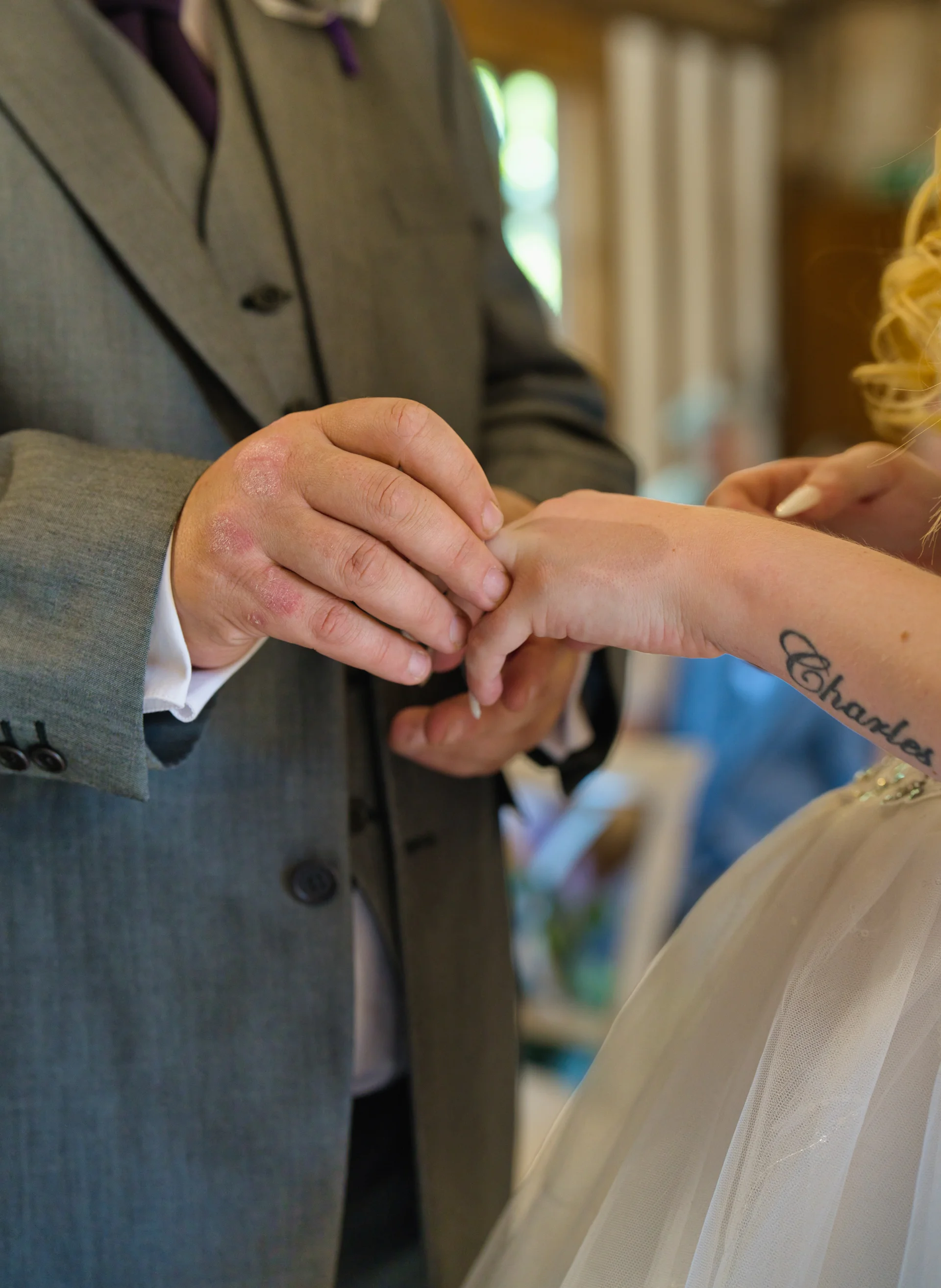 A close-up shot captures the groom in a grey three-piece suit gently placing a wedding ring onto the bride's finger, highlighting a wrist tattoo.