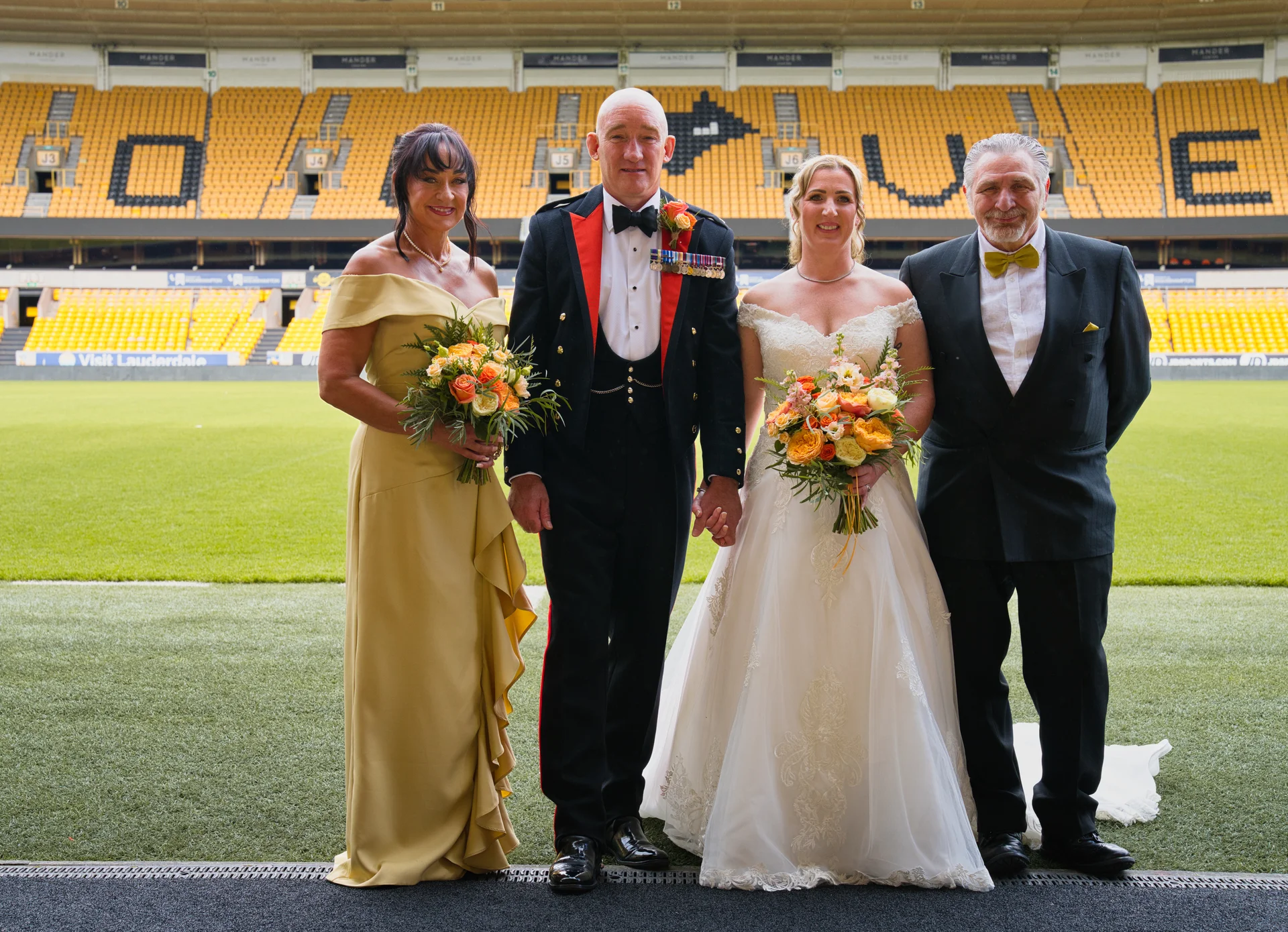 A bridal party including a bride in an off-the-shoulder gown, a groom in a military uniform, and two guests pose together holding bouquets inside a sports stadium.