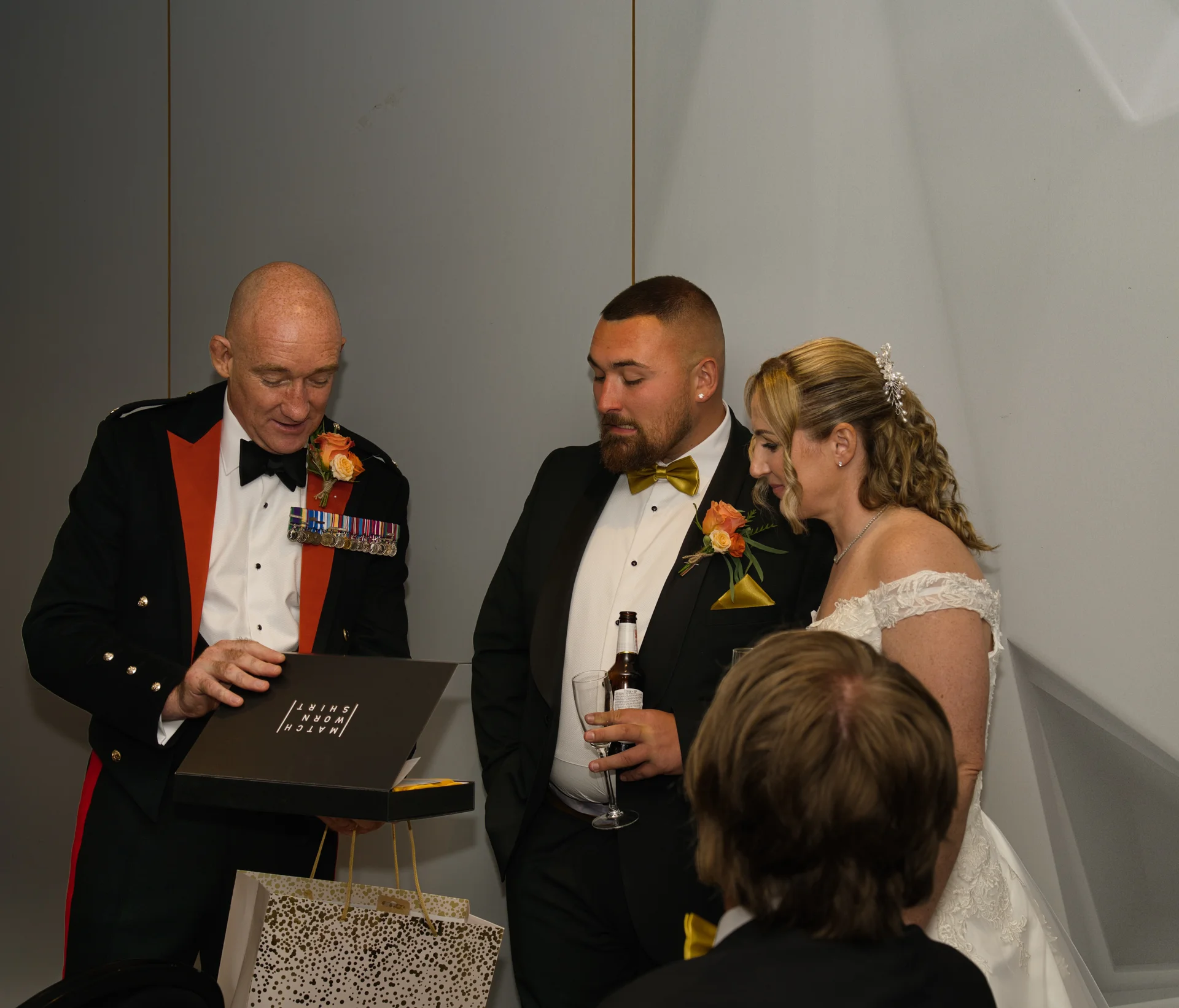 A newlywed couple and a guest look on as a man in a military uniform opens a presentation box during the wedding reception.