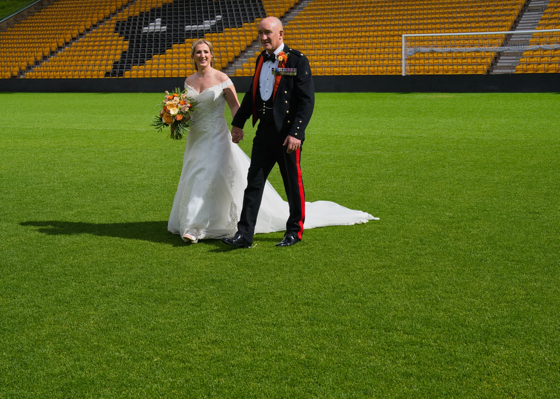 A bride in an off-the-shoulder white gown holding a floral bouquet walks hand-in-hand with a groom in a military uniform across the bright green pitch of a stadium.