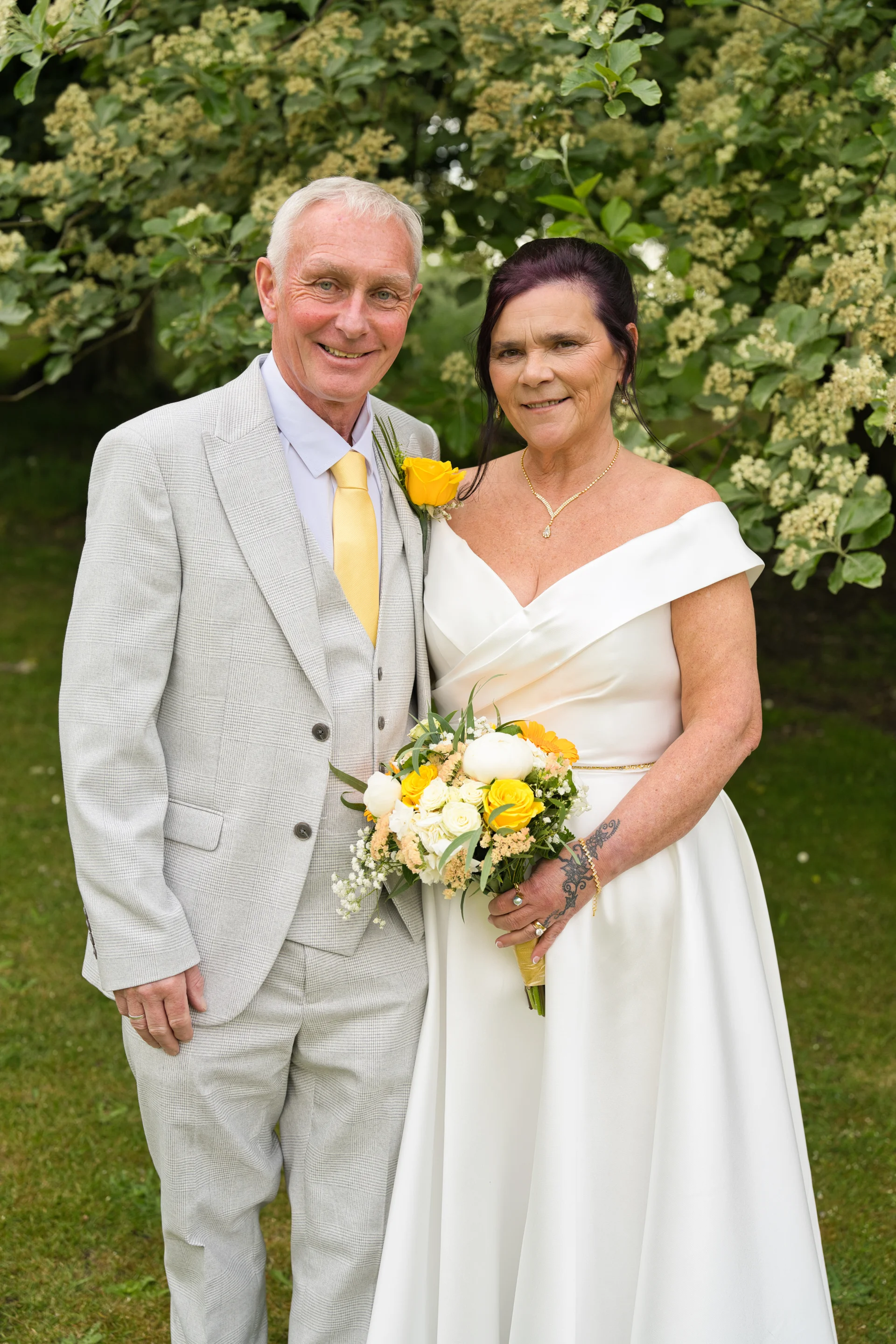An older couple smiling together outdoors in a garden setting, with the groom wearing a light grey suit and yellow tie, and the bride in an off-the-shoulder white satin gown holding a yellow and white bouquet.