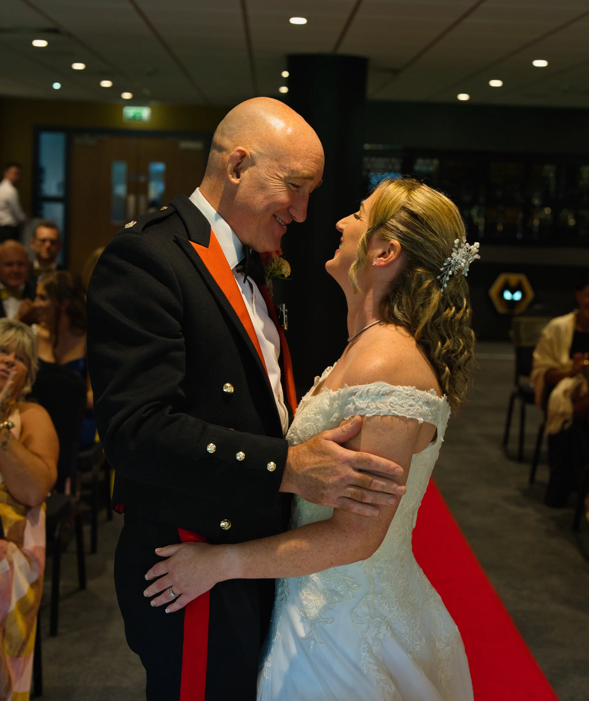 A smiling couple shares a loving embrace at their wedding, with the groom in a military dress uniform and the bride in an off-the-shoulder lace dress.