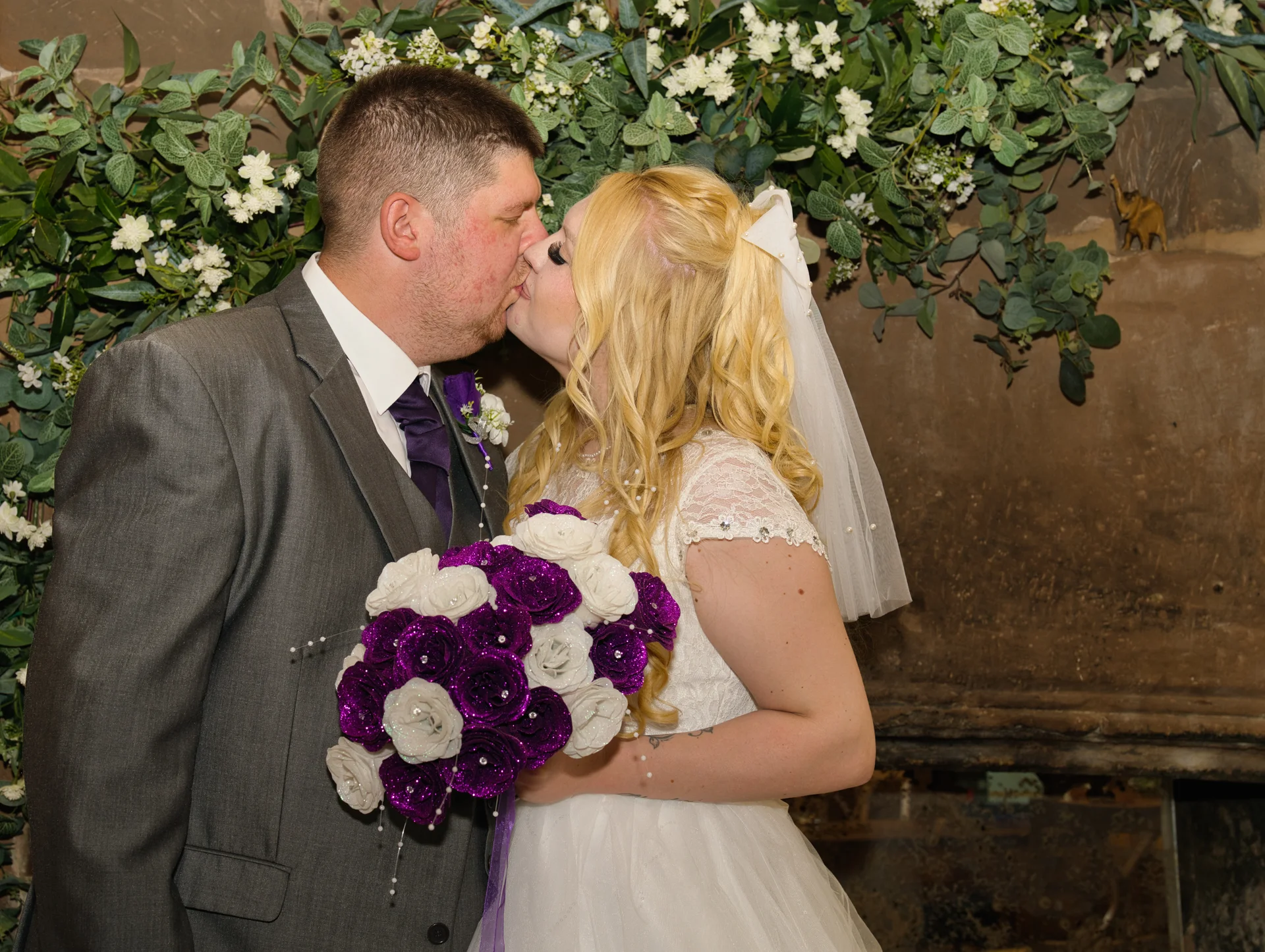 A newlywed couple shares a tender kiss against a stone wall adorned with hanging foliage and white flowers, with the bride holding a striking purple and white bouquet.