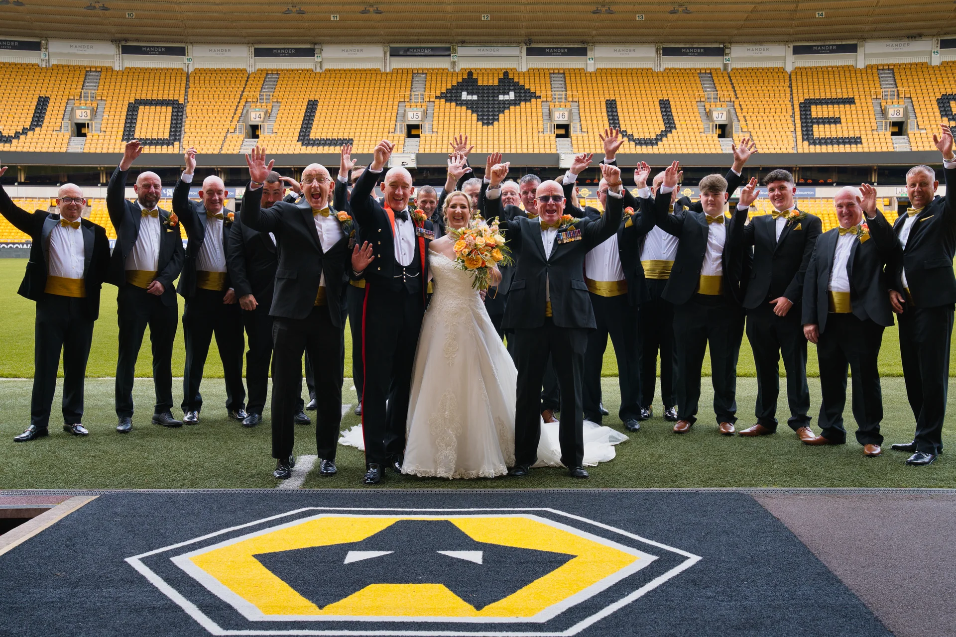 A bride and groom stand smiling with a large group of groomsmen raising their hands in celebration on the pitch of a football stadium.