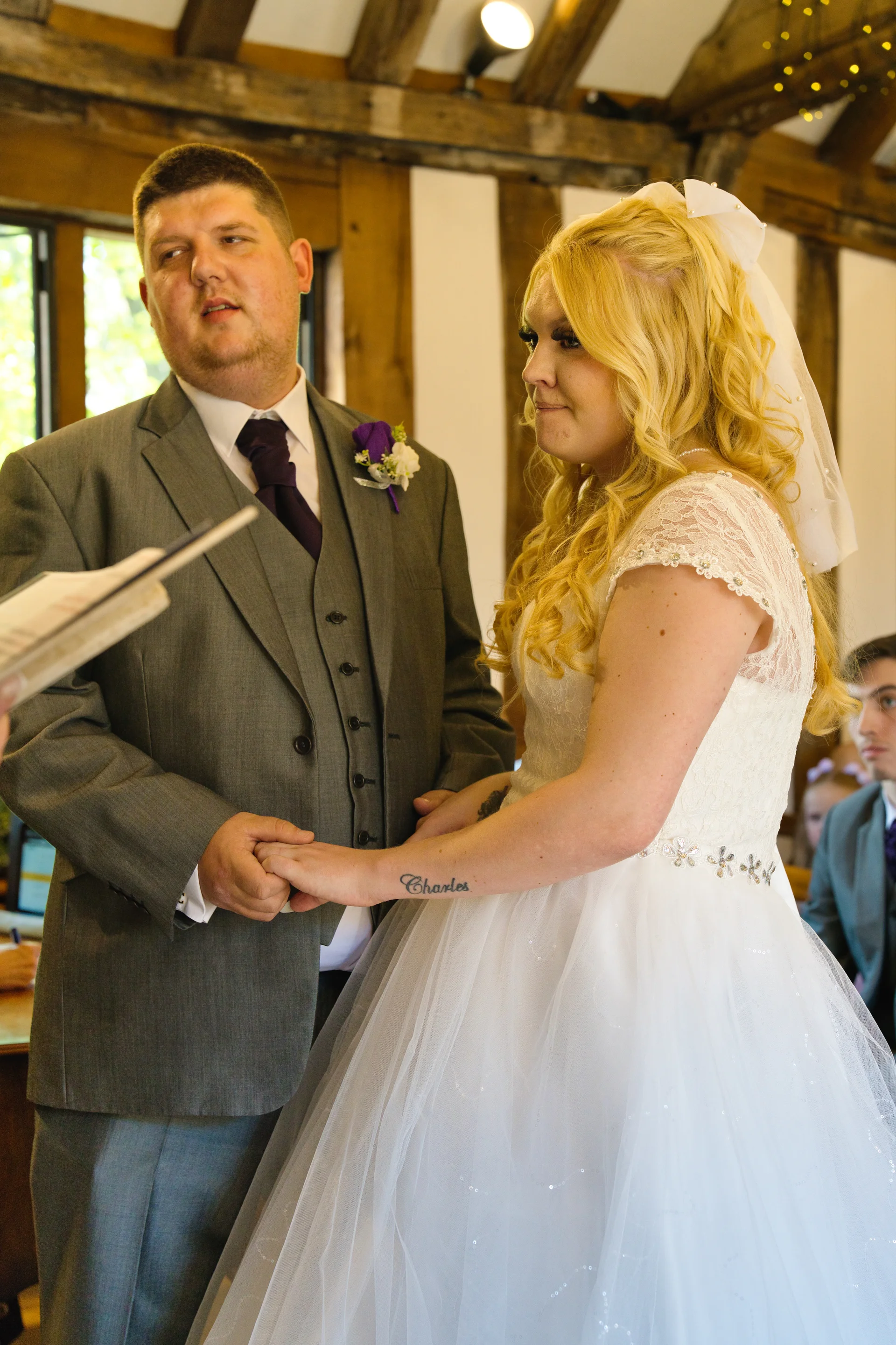 A bride and groom hold hands during their wedding ceremony inside a rustic barn venue, with the bride wearing a lace-bodice a-line dress and the groom in a grey three-piece suit.