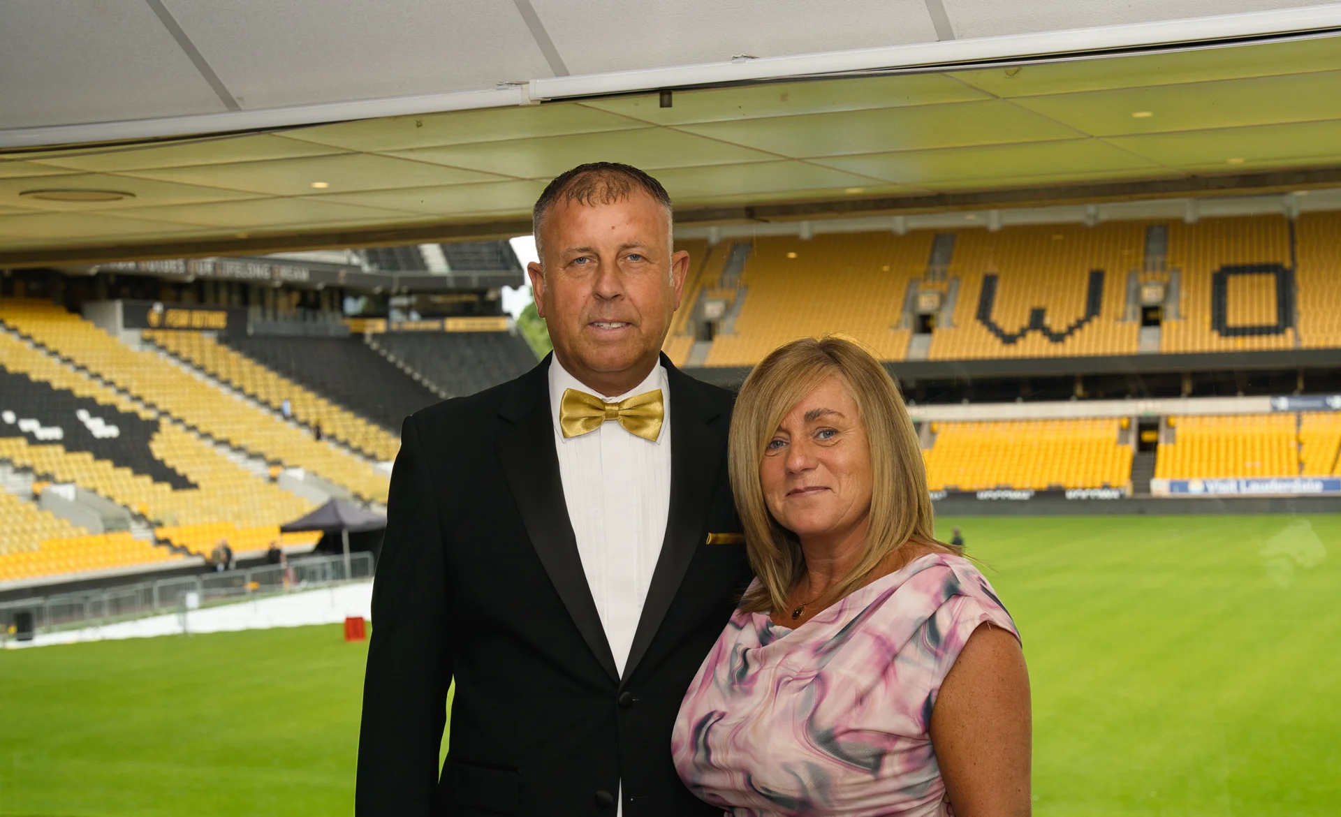 An older couple poses for a portrait inside a sports stadium wedding venue, with the man wearing a black tuxedo with a gold bow tie and the woman in a patterned pink dress.
