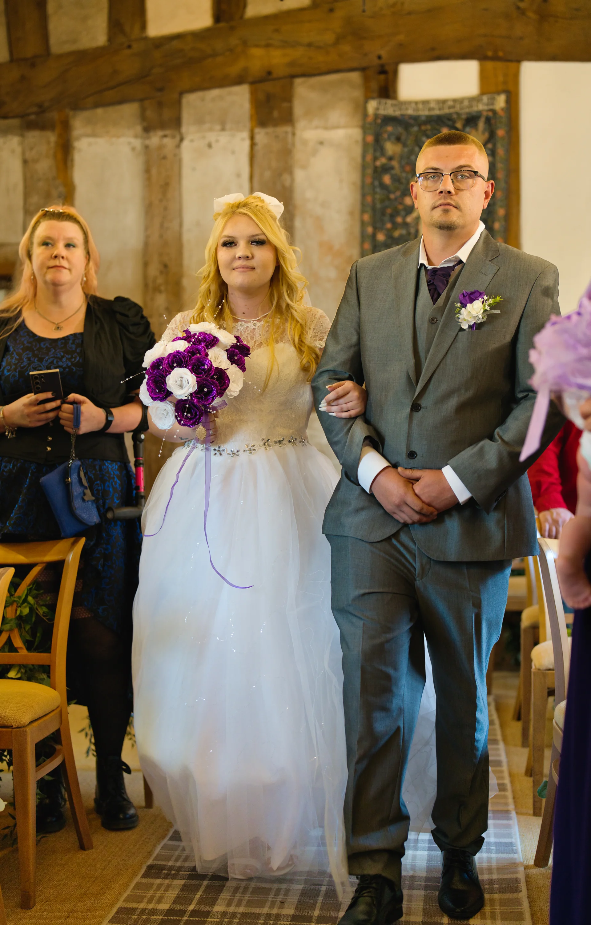 A bride and groom walk down the aisle together inside a rustic barn with stone walls, holding a purple and white bouquet.