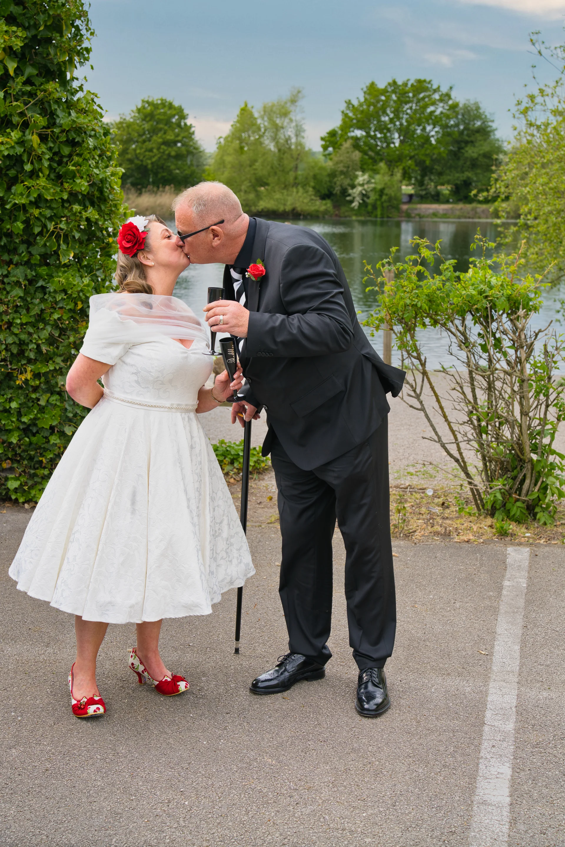 An older bride and groom share a tender kiss by a tranquil lake while holding champagne flutes and wearing coordinating red floral accessories.