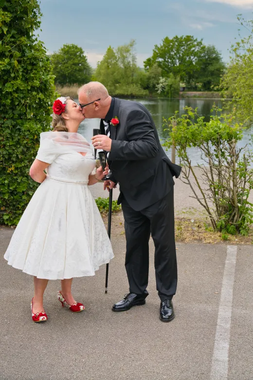 An older bride and groom share a tender kiss by a tranquil lake while holding champagne flutes and wearing coordinating red floral accessories.