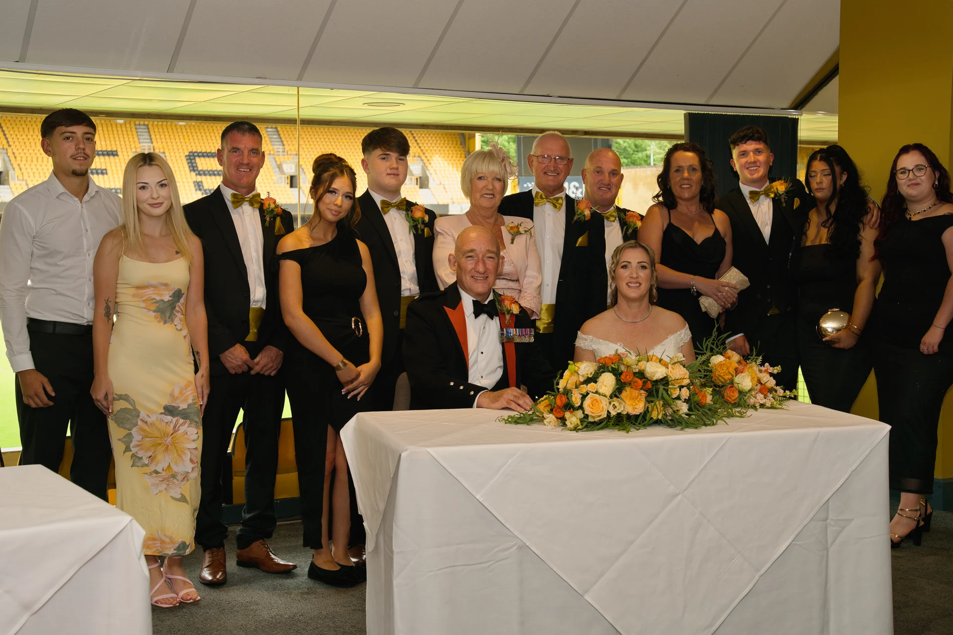 A large wedding party and family members pose together behind a beautifully decorated top table featuring orange and yellow floral arrangements inside a sports stadium venue.