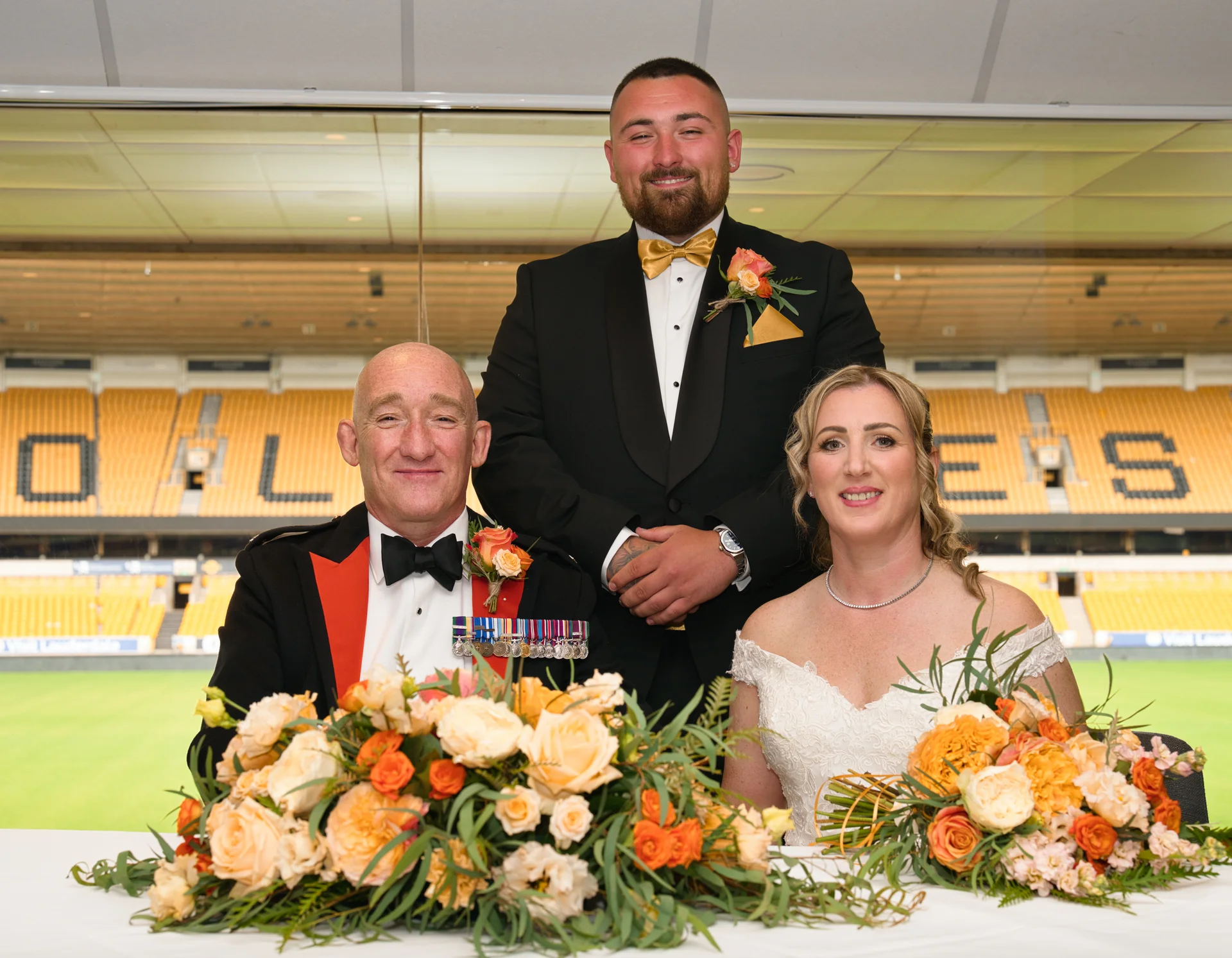 A newlywed couple and a guest in uniform pose together behind a floral-decorated table inside a sports stadium wedding venue.