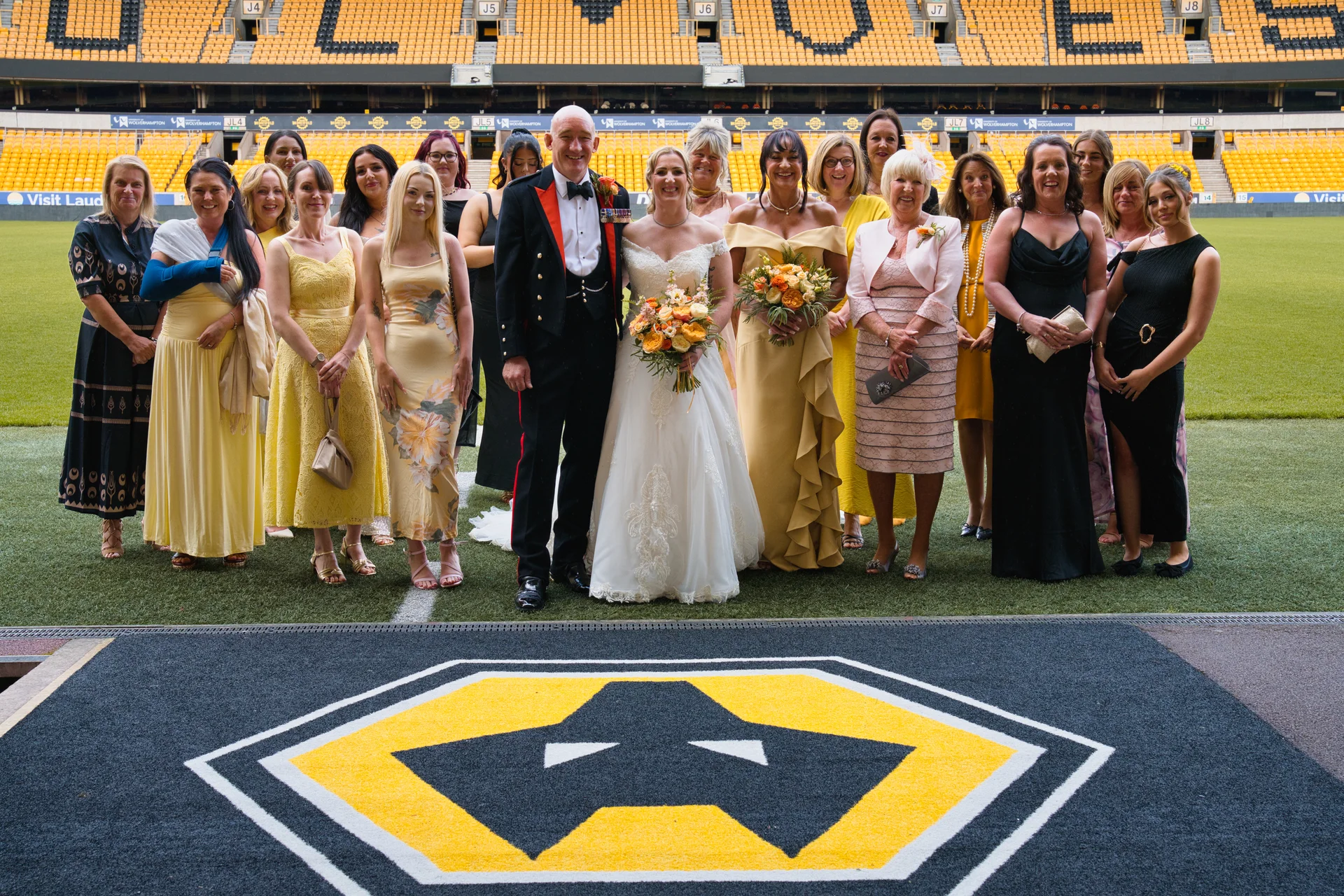 A newlywed couple posing with their large wedding party on the pitch of a sports stadium featuring yellow and black seating.