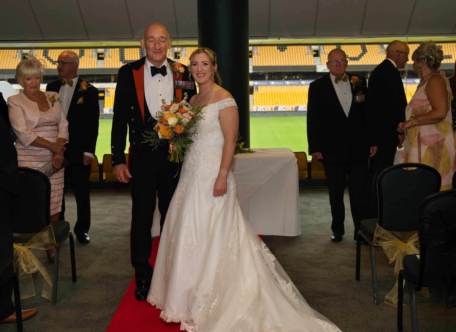 A smiling bride in an off-the-shoulder lace a-line gown and groom in military dress uniform stand on a red carpet inside a sports stadium wedding venue with guests watching.