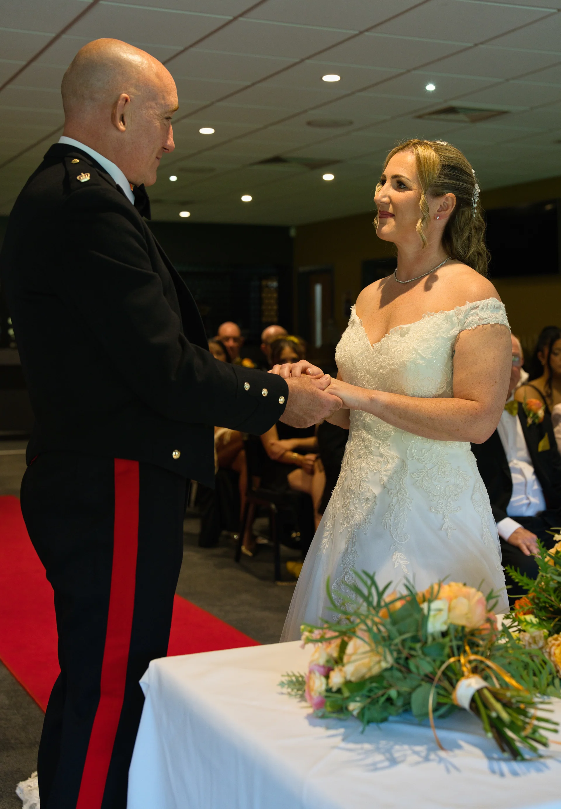 A groom in a military dress uniform holding hands and exchanging loving glances with his bride in an off-the-shoulder lace wedding gown during their indoor ceremony.