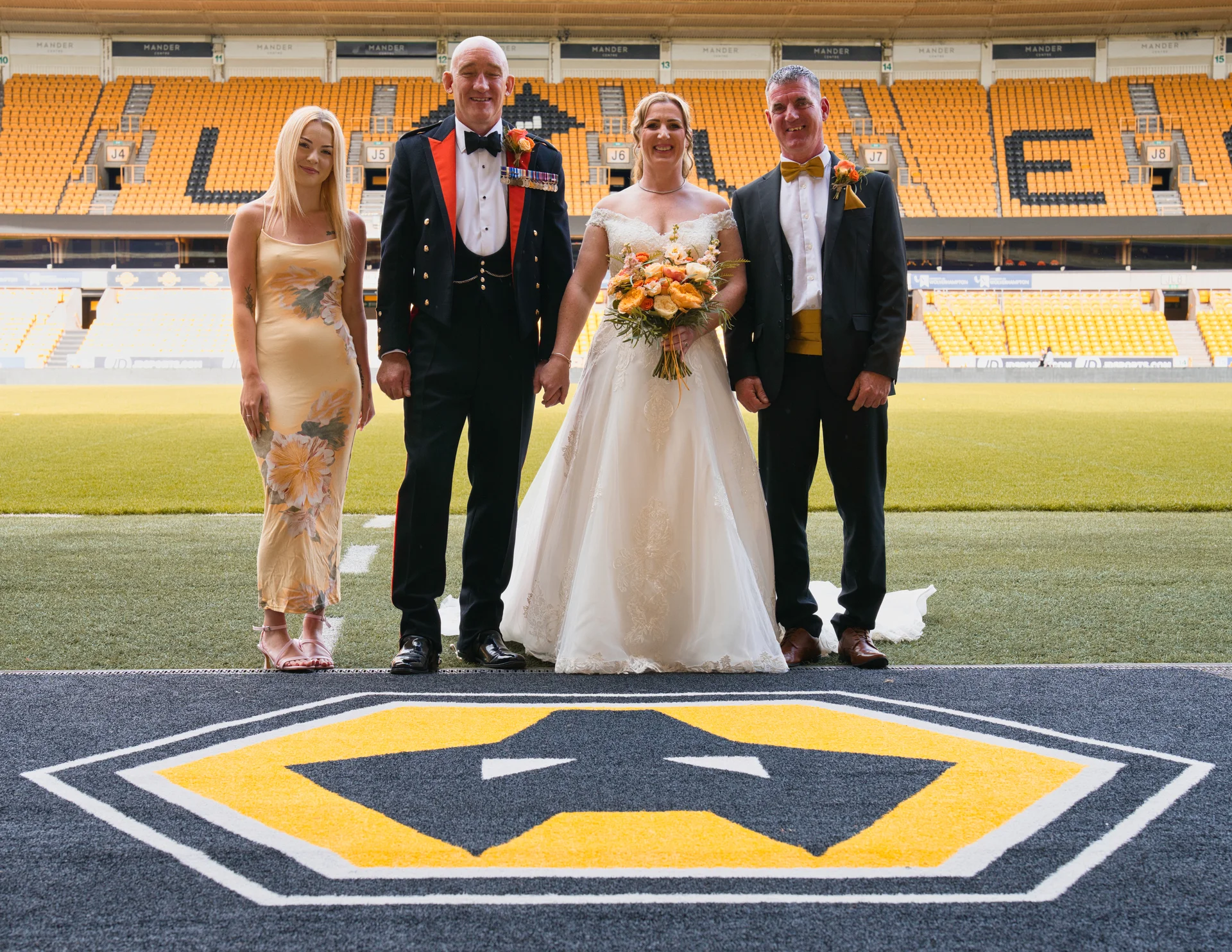 A bride in a white gown and groom in a military uniform stand alongside another couple on the pitch of a yellow-seated football stadium.