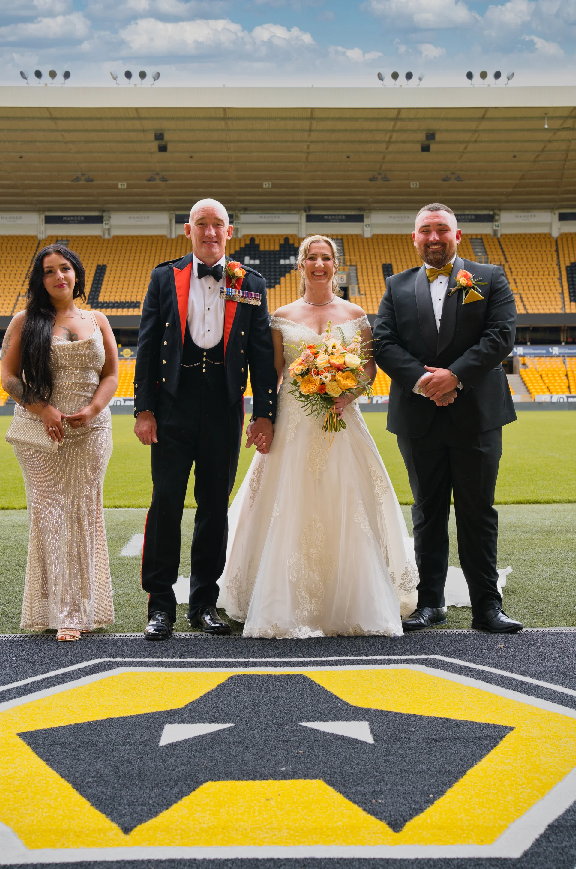 A bridal party poses formally on the pitch inside a large sports stadium with yellow seating in the background.
