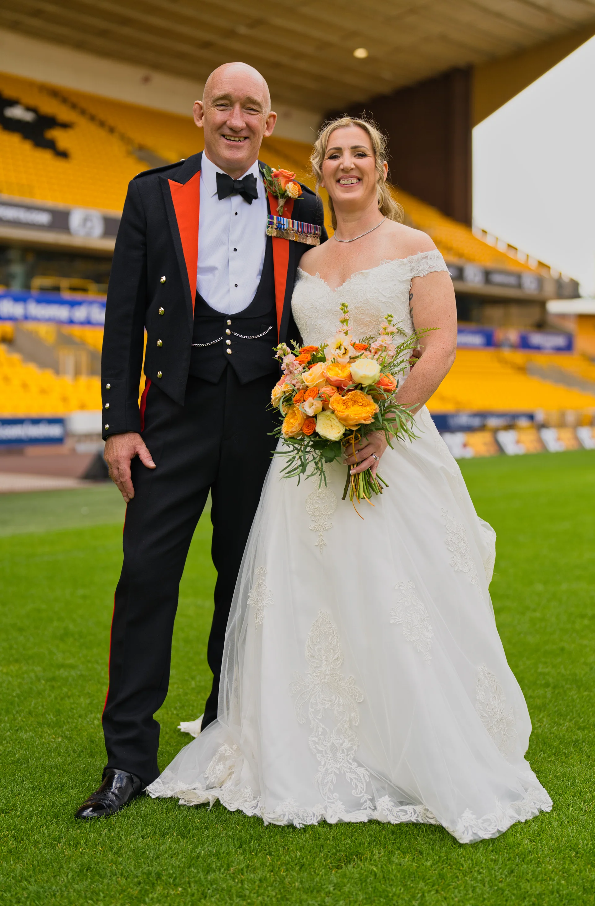 A smiling bride holding a vibrant orange and yellow floral bouquet poses alongside her groom in a military dress uniform on a sports stadium pitch.