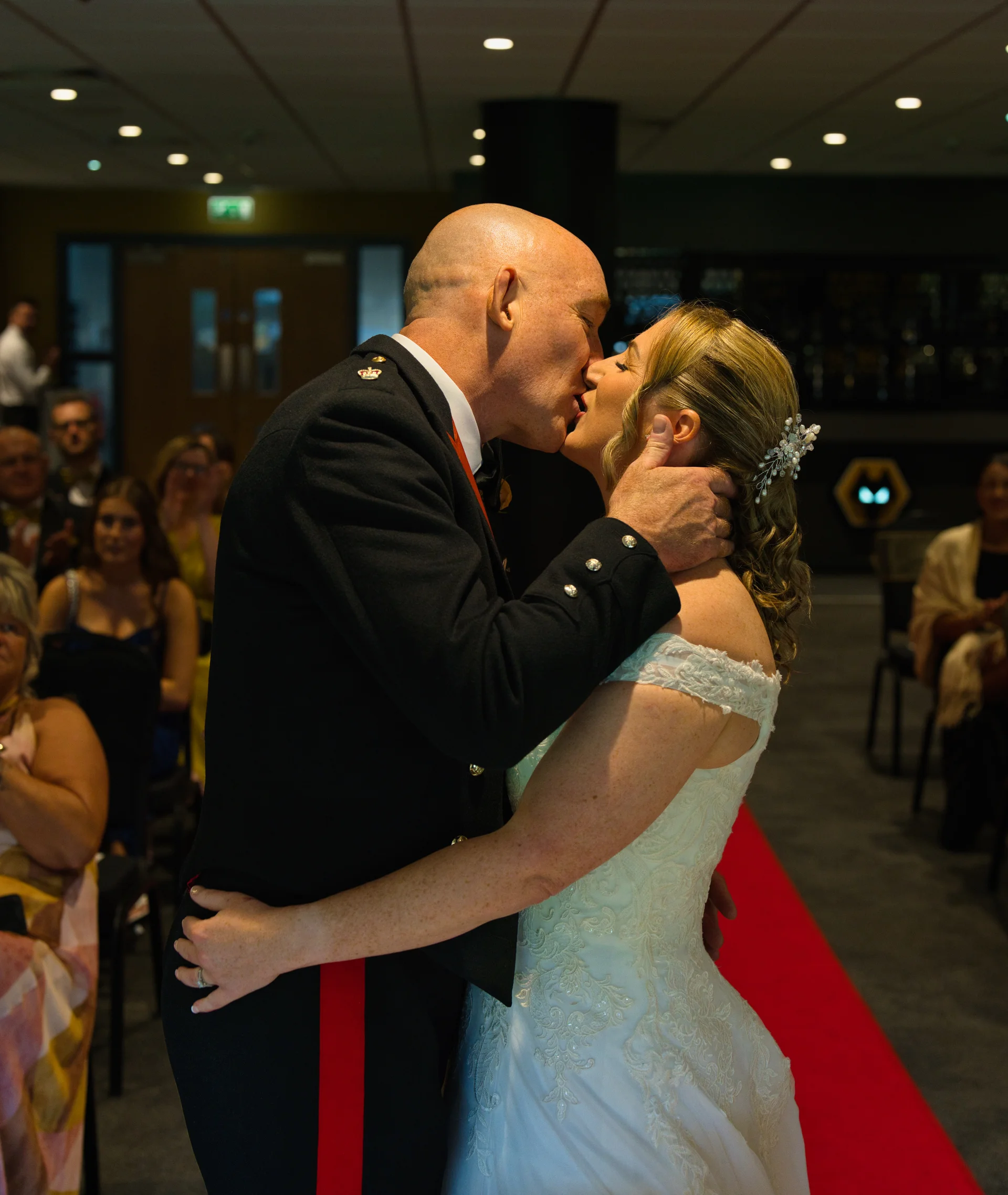 A groom in a military uniform shares a passionate first kiss with his bride wearing an off-the-shoulder lace gown as wedding guests watch on.