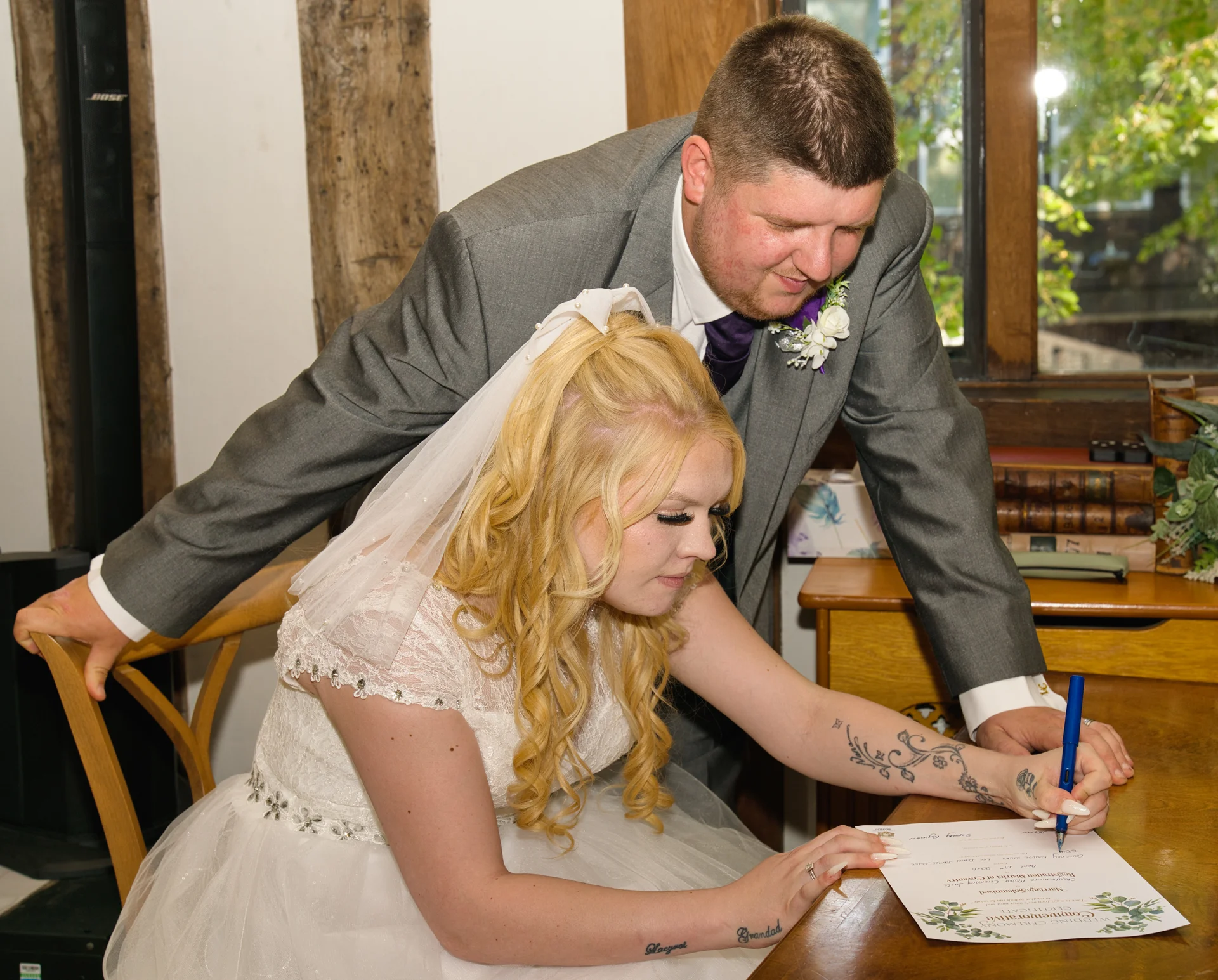The bride signs the wedding certificate at a wooden table while the groom in a grey suit leans over to watch.