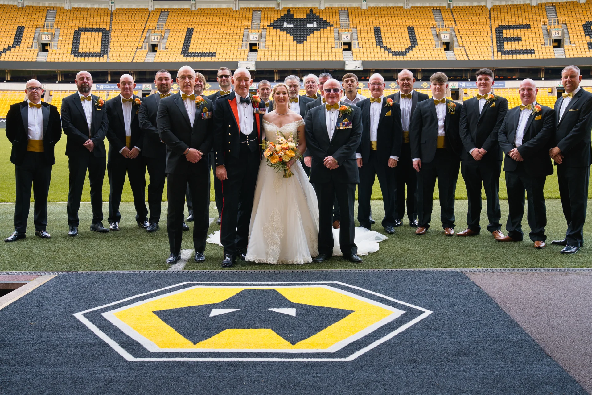 A newlywed couple poses with a large group of groomsmen wearing black tuxedos on the pitch of a sports stadium.
