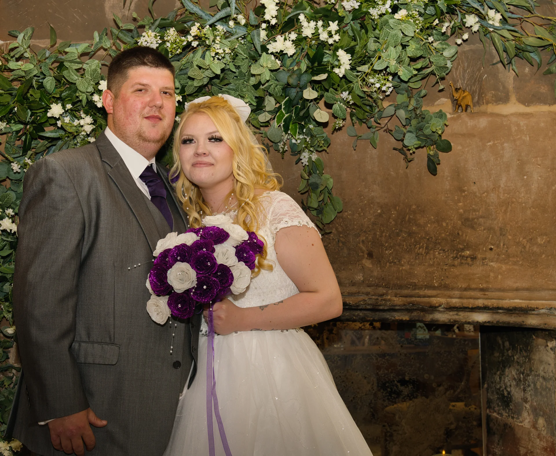 A newlywed couple poses together in front of a large stone fireplace adorned with lush green foliage and white flowers, with the bride holding a purple and white rose bouquet.