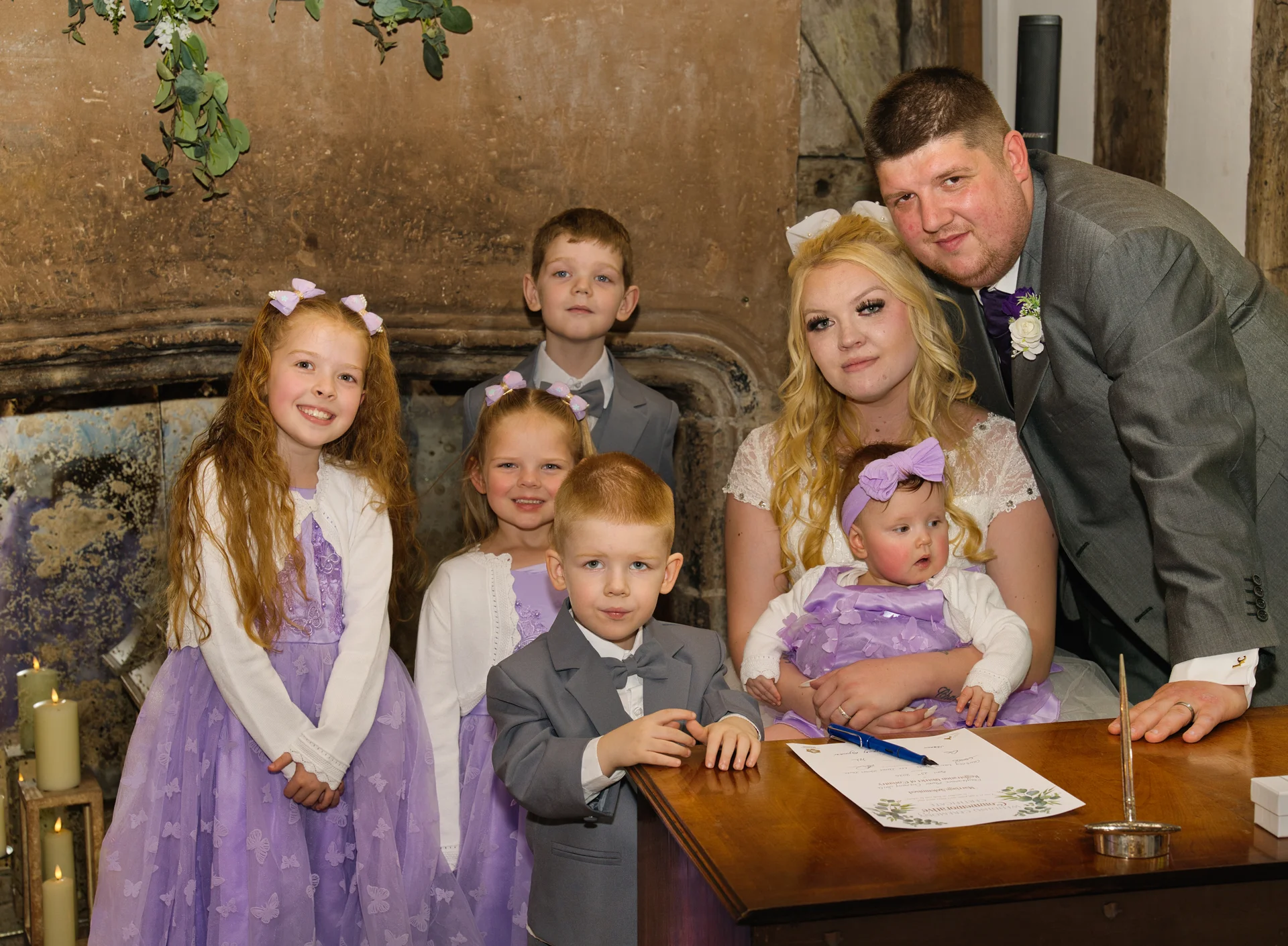 A family gathers around a wooden signing table with a bride and groom in a rustic venue featuring a large stone fireplace.