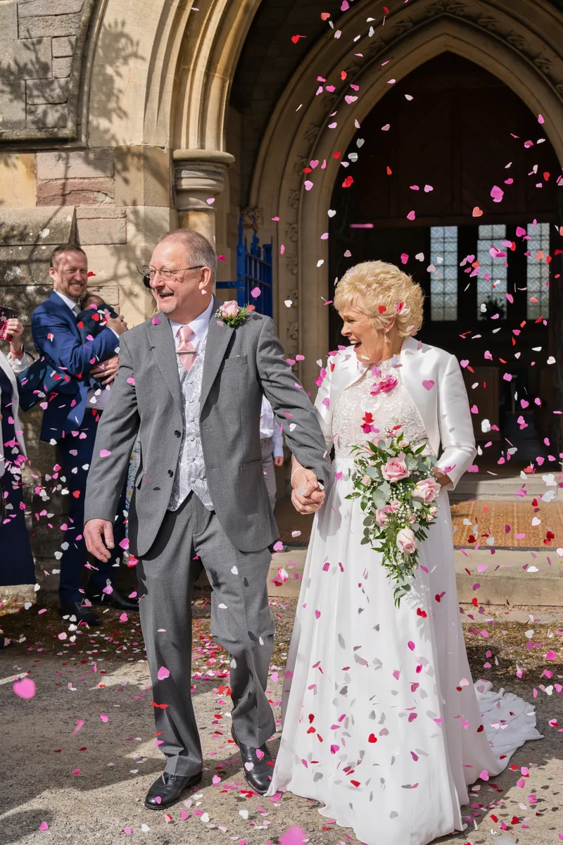 An older couple joyfully exits a stone church as pink heart-shaped confetti cascades around them hand in hand.