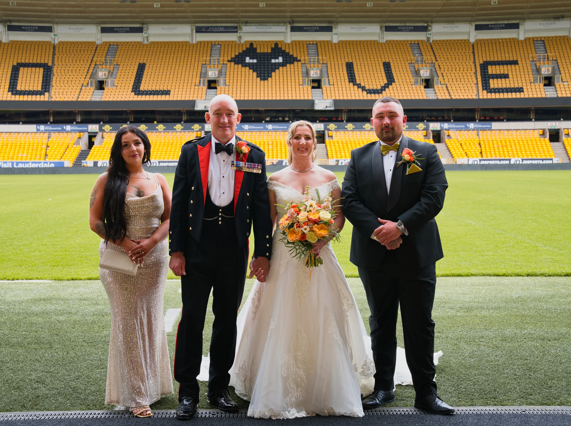 A newly married couple in wedding attire and military dress pose alongside two wedding guests on the pitch of a large football stadium with yellow seating.