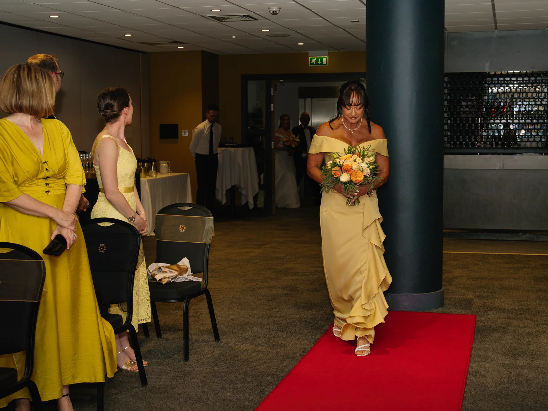 A woman in an off-the-shoulder yellow dress holding a bouquet walks down a red carpet as guests look on in a hotel ballroom.