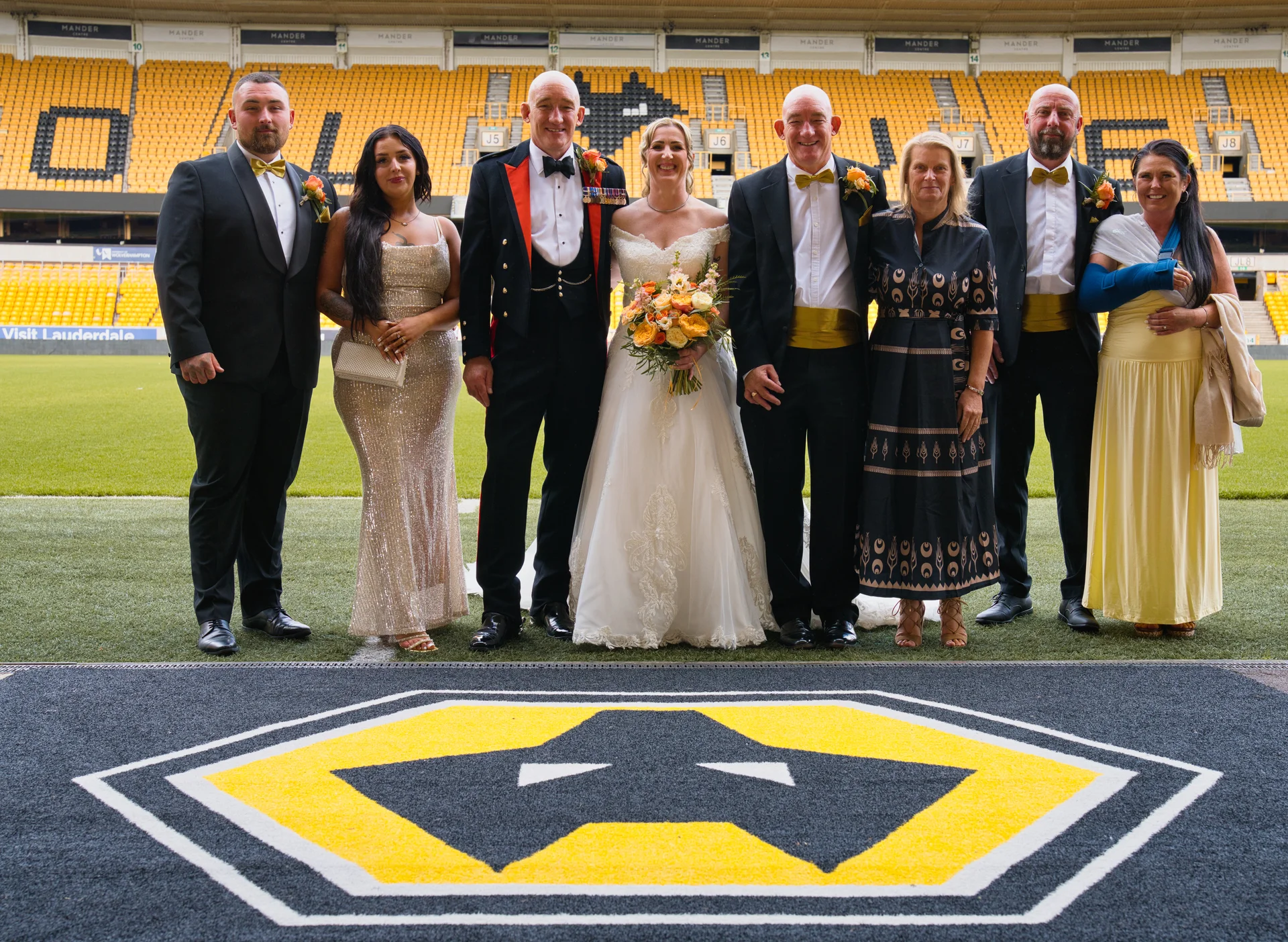 The bride and groom pose with their wedding party on the pitch inside a football stadium, standing proudly in front of the stands.