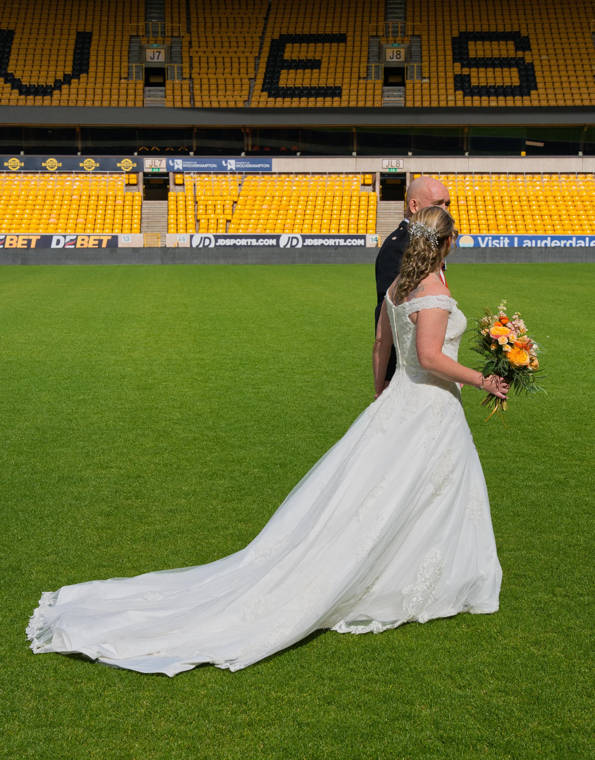 A bride in an off-the-shoulder lace a-line gown with a long train and groom walk across the green pitch of a large sports stadium carrying a bright bouquet.
