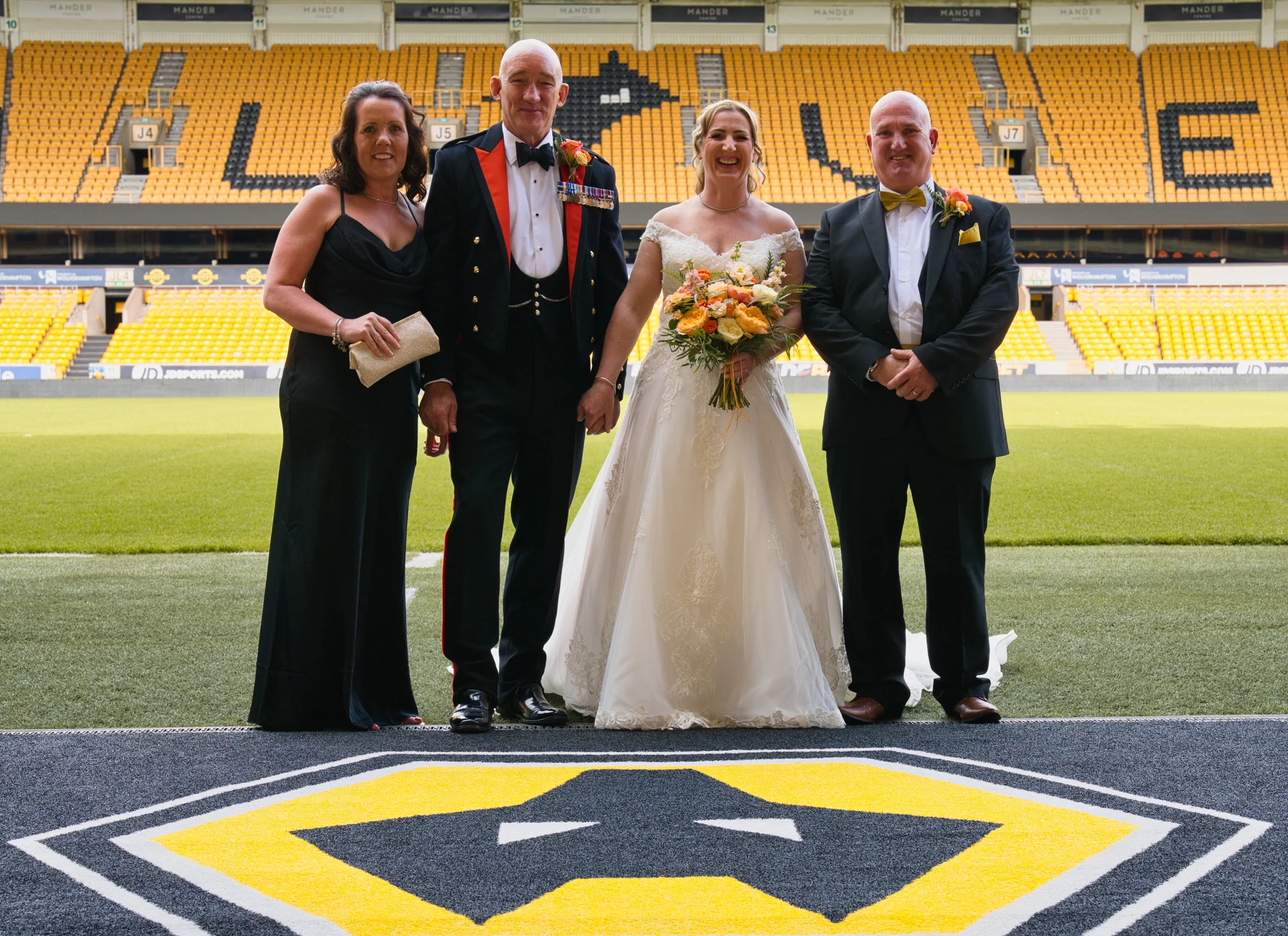 A bride in an off-the-shoulder lace a-line gown holding a bouquet stands alongside family members inside a football stadium.